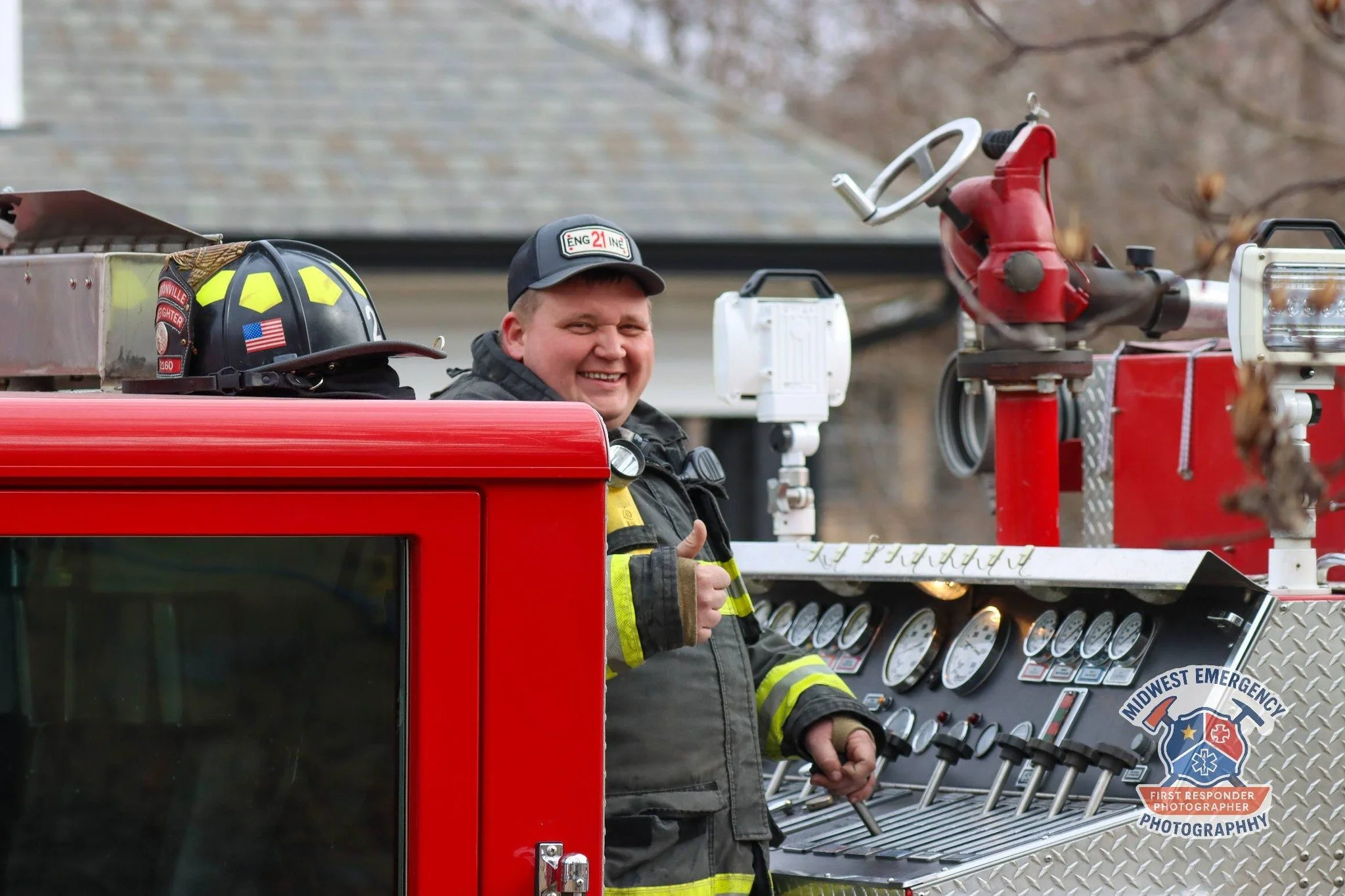 A smiling firefighter gives a thumbs-up while standing on a fire truck, wearing a uniform and helmet, with fire fighting equipment and gauges visible on the vehicle.