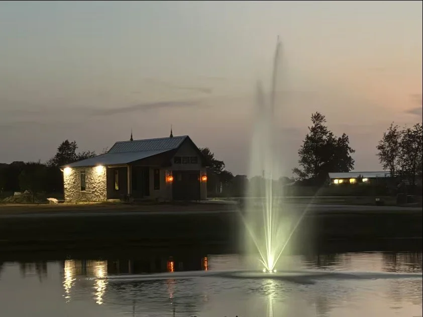 A pond with a lit water fountain in the foreground, and a small house with bright exterior lighting and trees in the background during dusk.