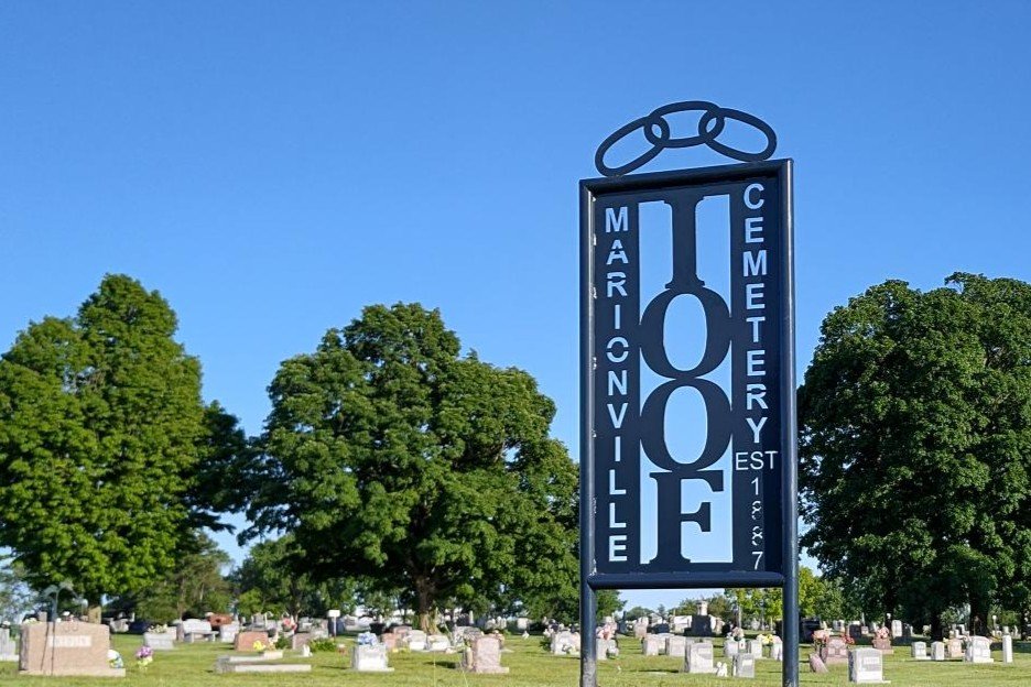 Sign for the Coffee Cemetery in Marionville, established in 1907, with a cemetery in the background and trees under a clear blue sky.