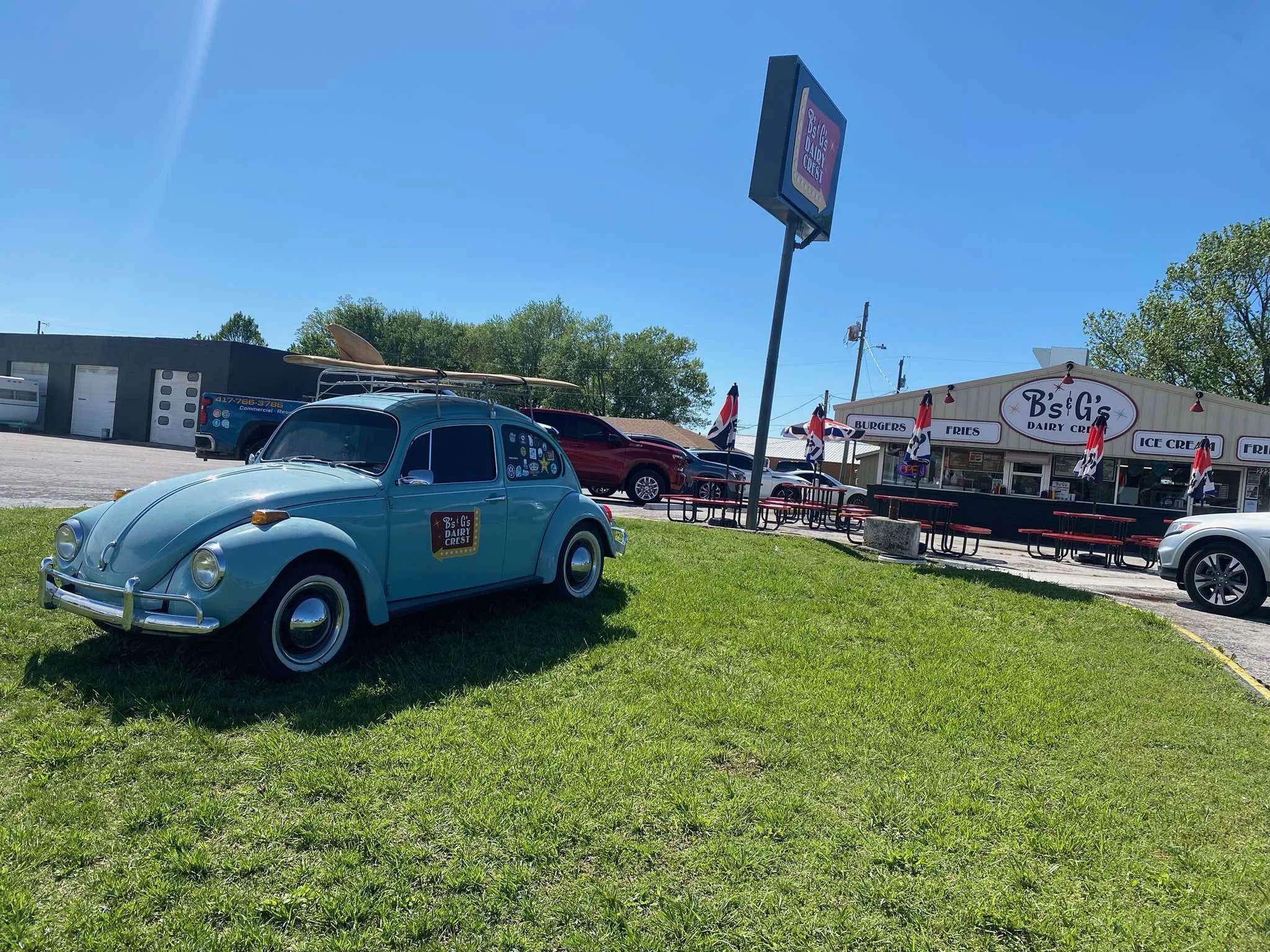 A vintage blue Volkswagen Beetle with a surfboard on the roof parked on a grassy area outside B's G's Dairy Crust restaurant, with outdoor seating and flags in the background.