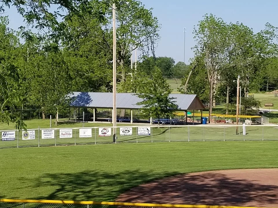 A baseball field with a green grassy outfield, a dirt infield, and a covered pavilion in the background. There are trees around the field and advertising signs along the outfield fence.
