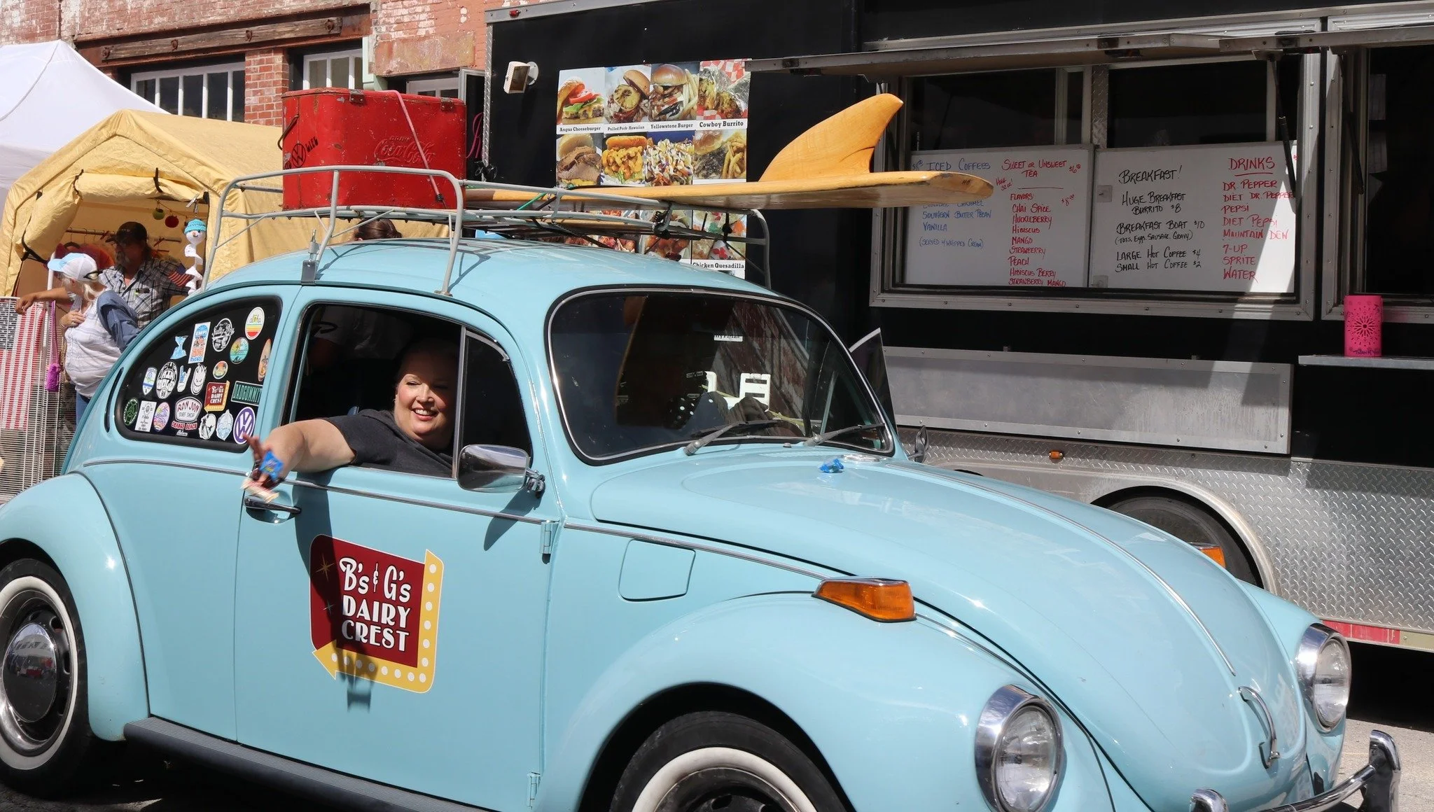 A vintage light blue Volkswagen beetle with stickers on the window, parked in front of a food truck with a person inside giving a thumbs up. The food truck displays a menu and images of burgers and breakfast items.