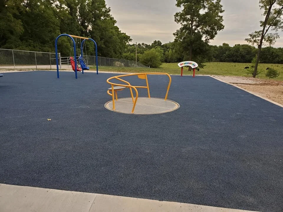 Empty playground with a blue rubber surface, a yellow circular spinning ride, a blue and red swing set, and a colorful seesaw in a park with trees and greenery in the background.