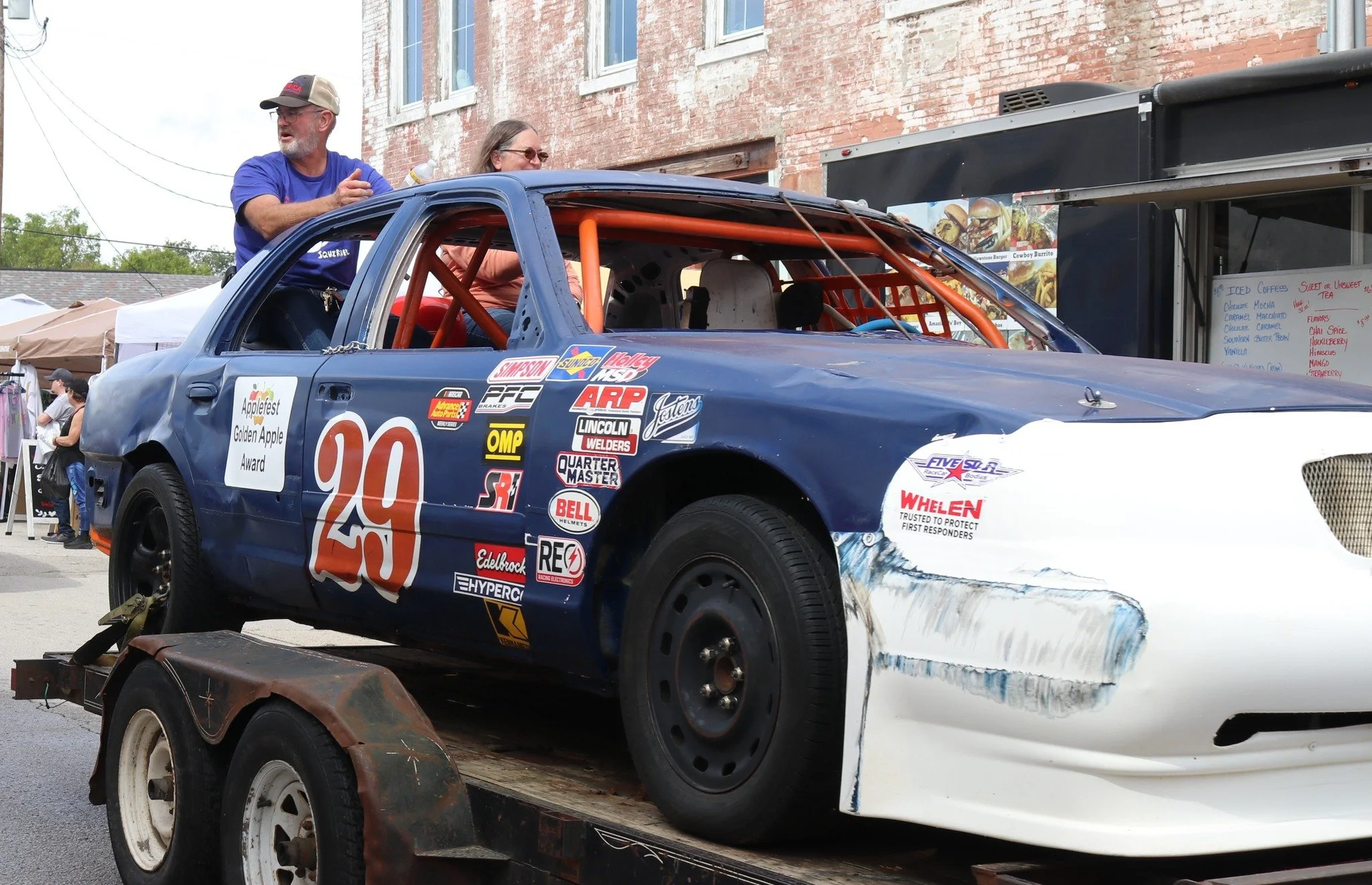A race car with the number 29 on the side, on a flatbed trailer in an outdoor setting. The car is blue with various sponsor stickers and has visible wheel and body damage. Two people are standing on the car's roof, talking.