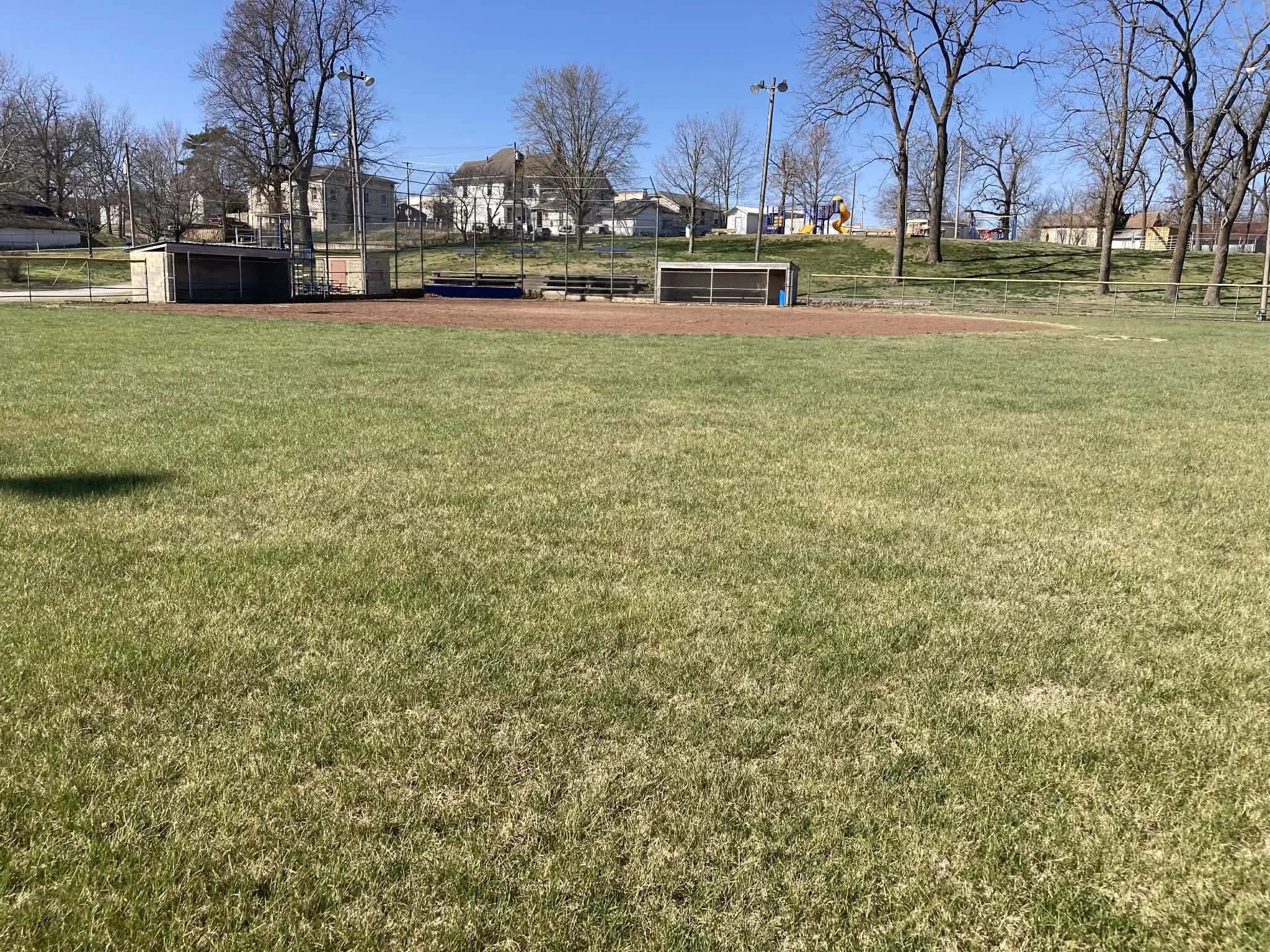 Empty baseball or softball field with green grass, dirt infield, and backstop, surrounded by a fence and residential houses in the background on a clear, sunny day, at Marionville, MO City park.