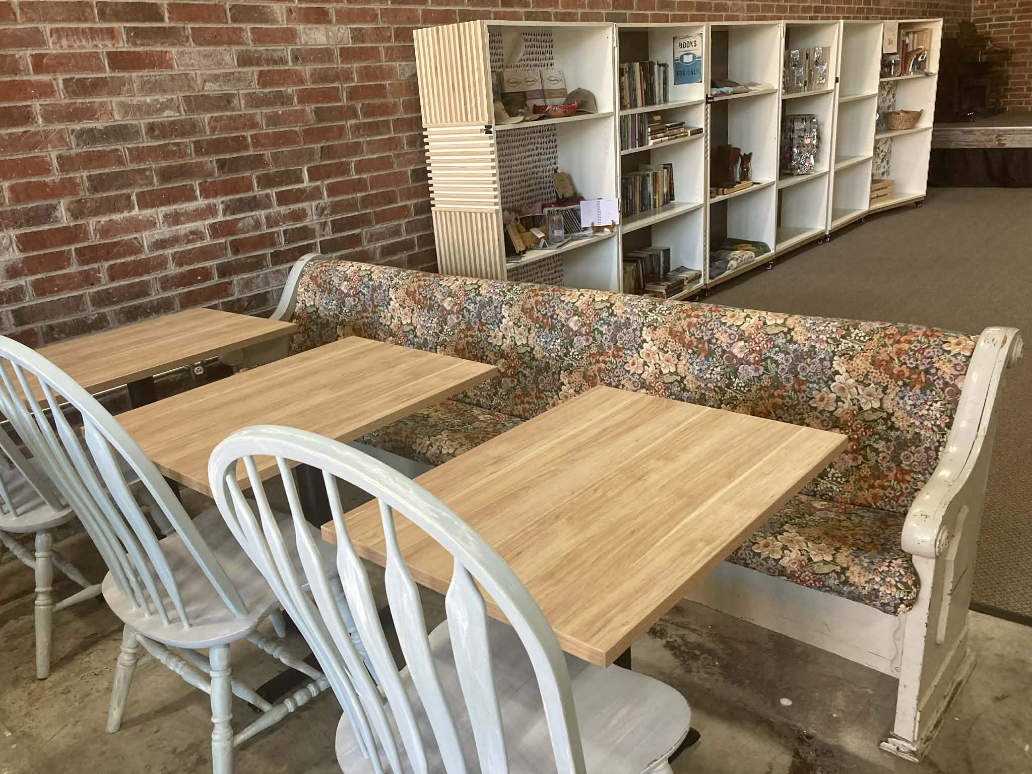 A cozy corner in a cafe or library with a brick wall, a floral patterned booth seating, wooden tables, white chairs, and a bookshelf filled with books and decorative items.