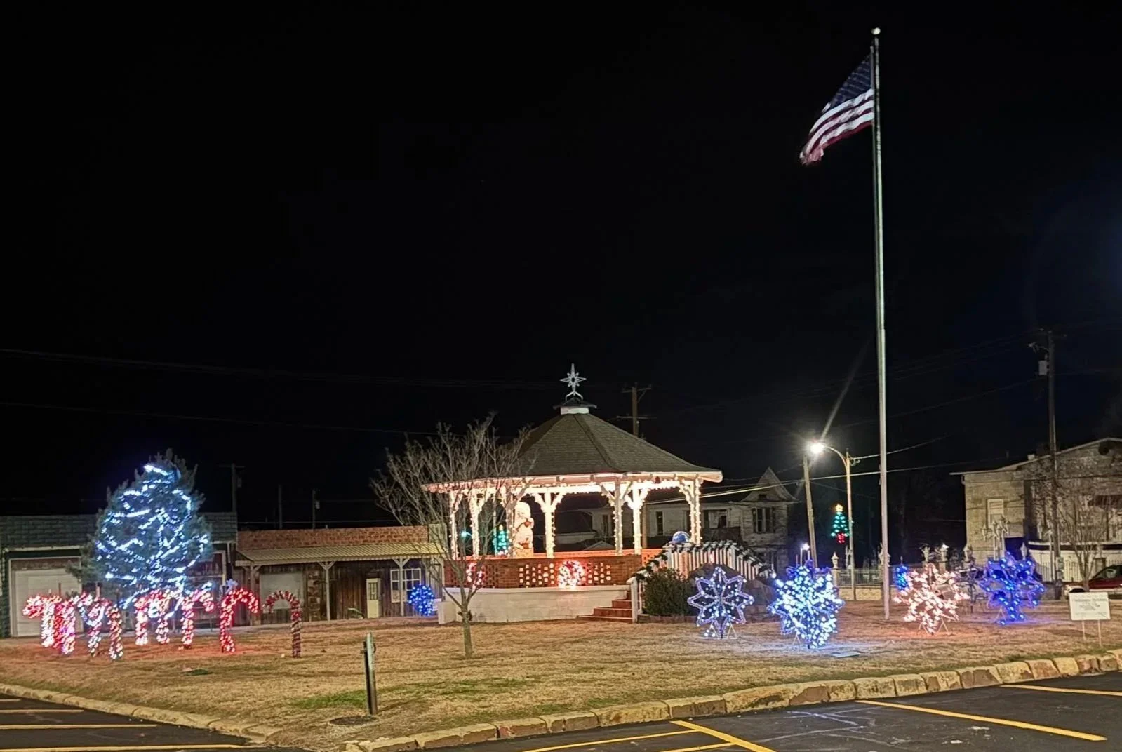 A decorated park at night with festive holiday lights, a pavilion with a star on top, a tall American flag, and illuminated snowflake and Christmas light displays.