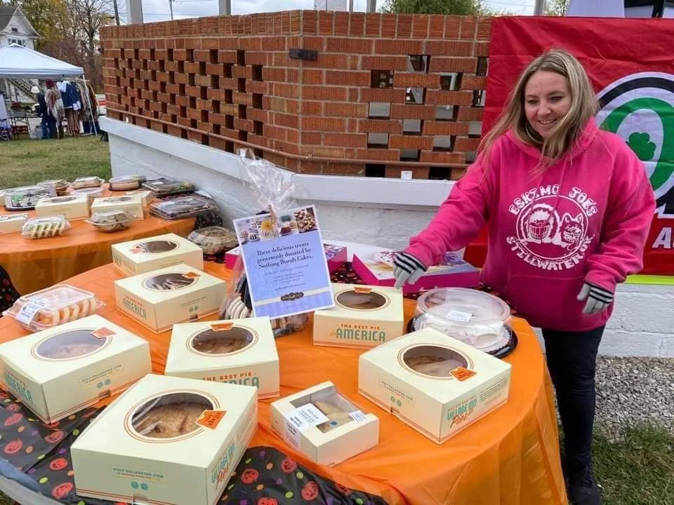 A woman in a pink hoodie standing at a table with boxed pies, at an outdoor market or fair.