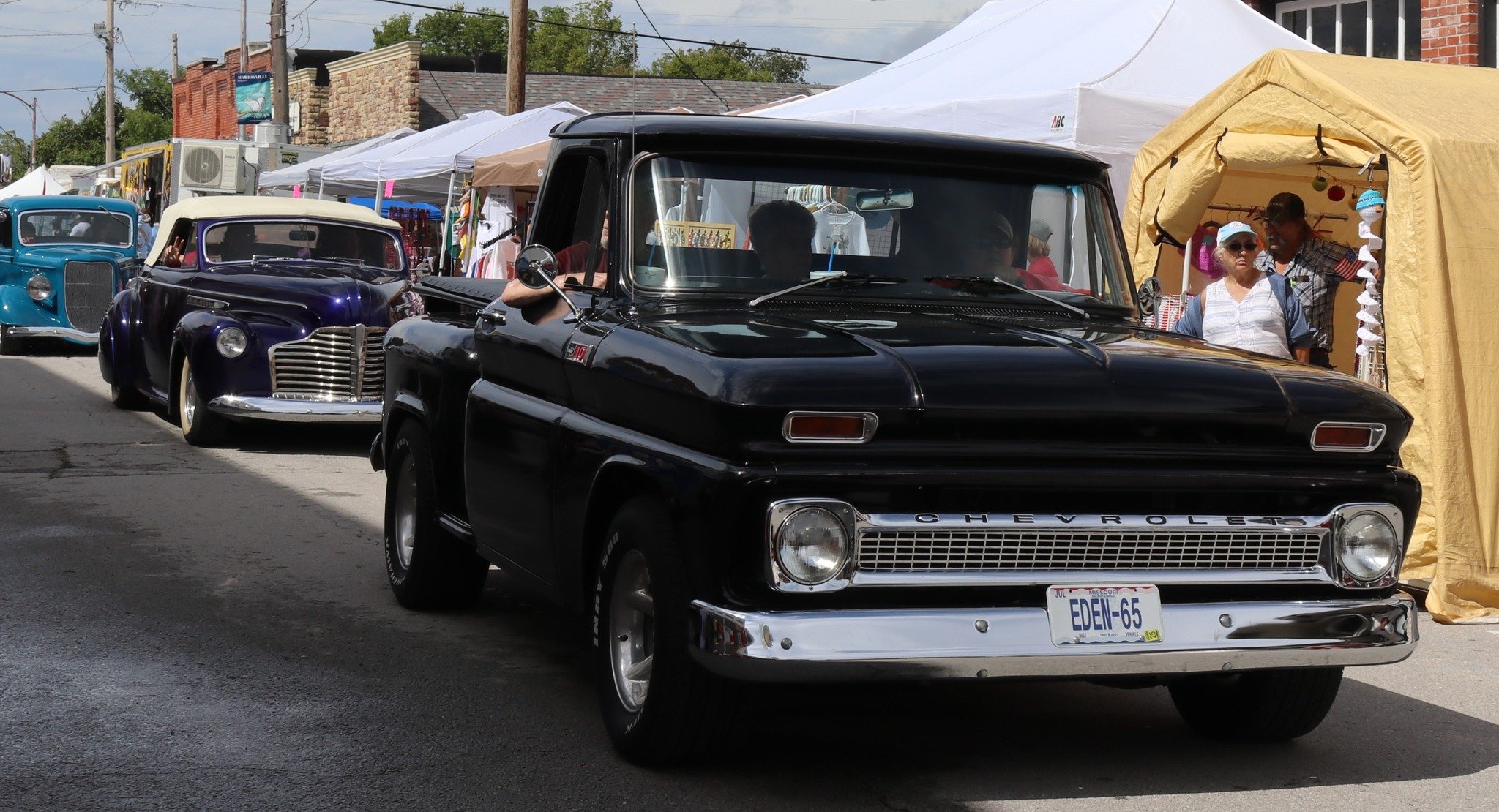 A line of vintage cars, including a black Chevrolet pickup truck in the foreground, parked along a street at a car show or festival, with tents and people in the background.