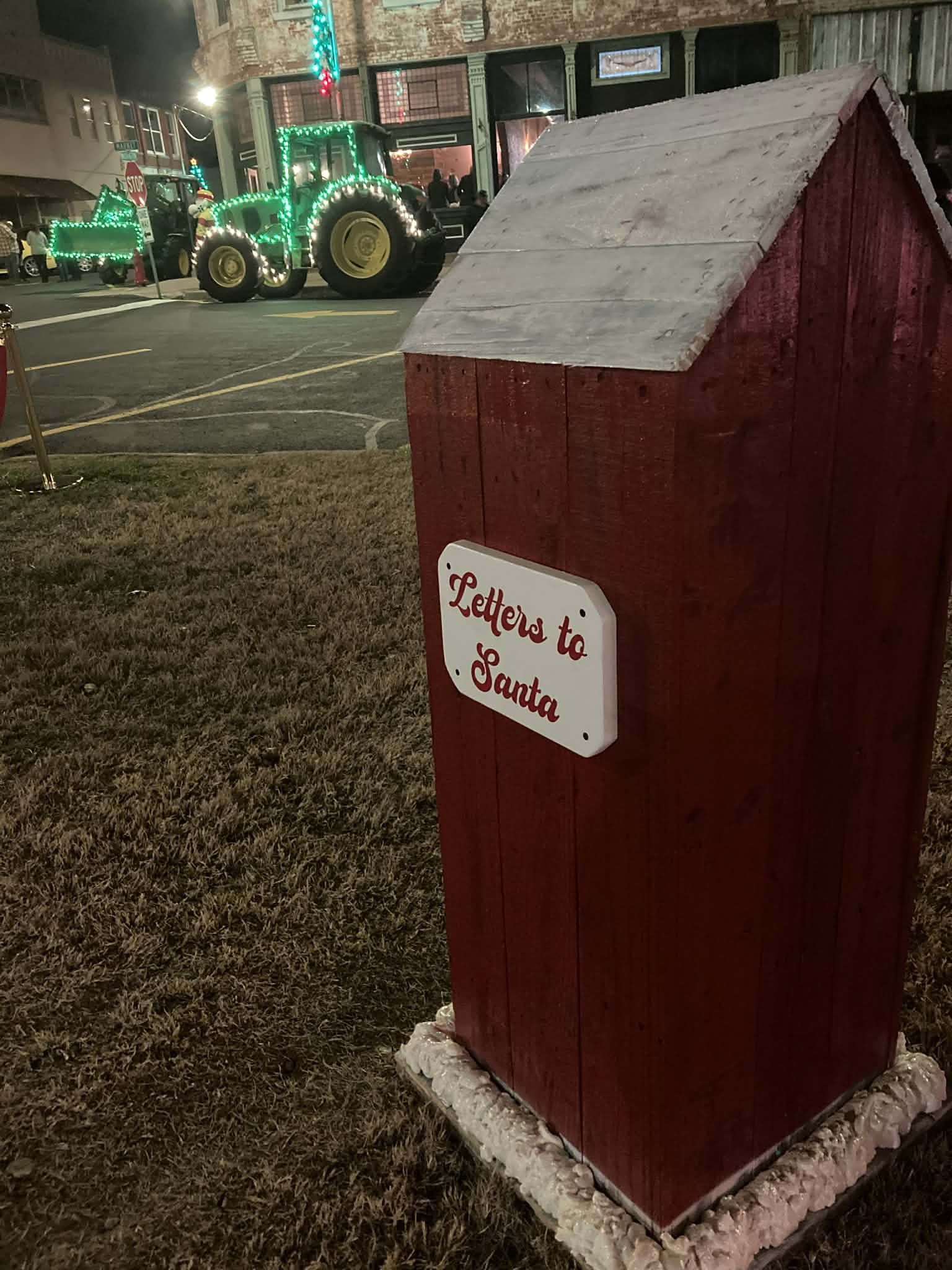 Red mailbox with a white sign reading 'Letters to Santa' on a patch of grass at night. In the background, a tractor decorated with green and red Christmas lights and a Christmas float are visible.