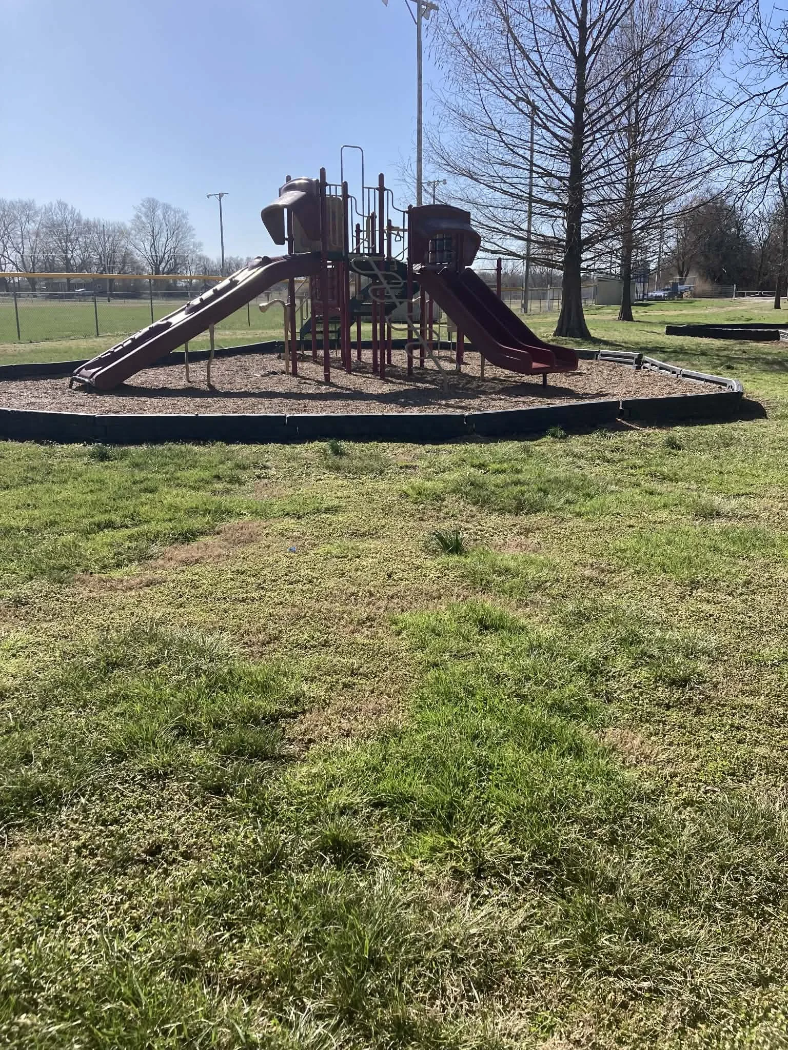 Empty playground with two slides, and climbing structures surrounded by trees on a sunny day. Marionville, MO city park.