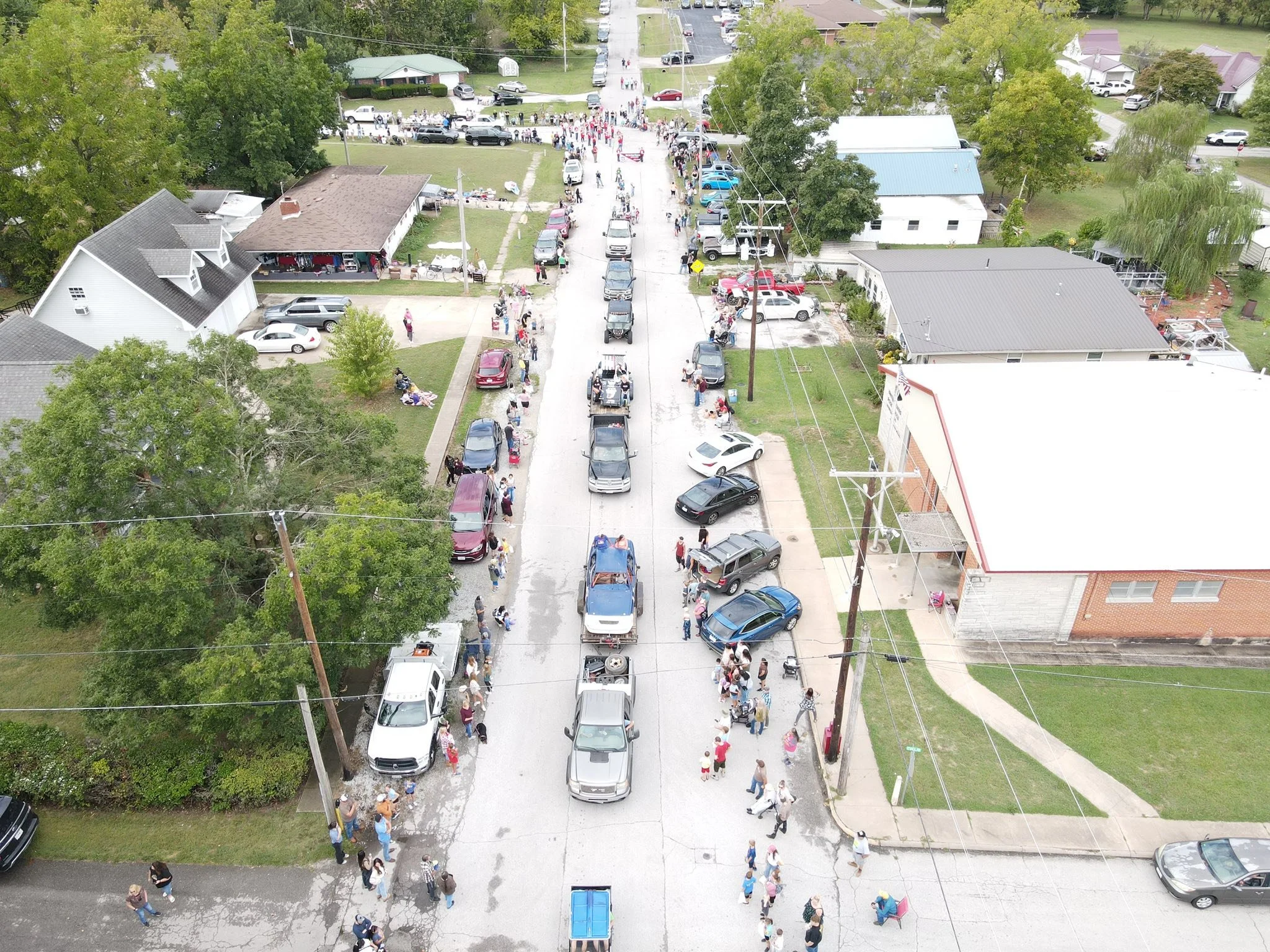 Bird's eye view of a street fair with people walking along the sidewalk, parked cars on both sides, and a line of vehicles in the street. Residential houses and trees are visible in the background.