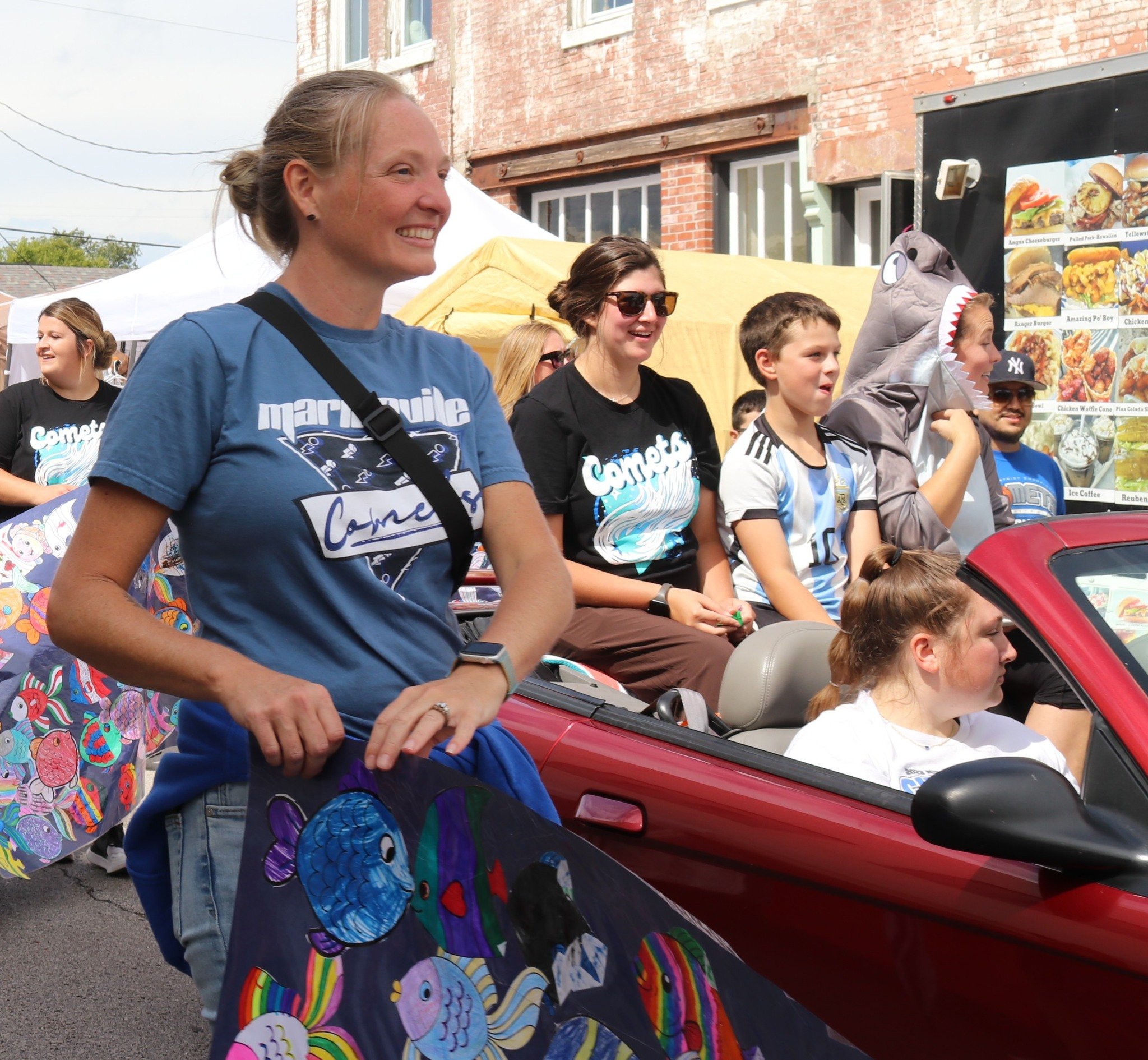 People participating in a parade; a woman in a blue Marquette t-shirt smiling, carrying a colorful fish-themed banner, and sitting in a red convertible car with children.