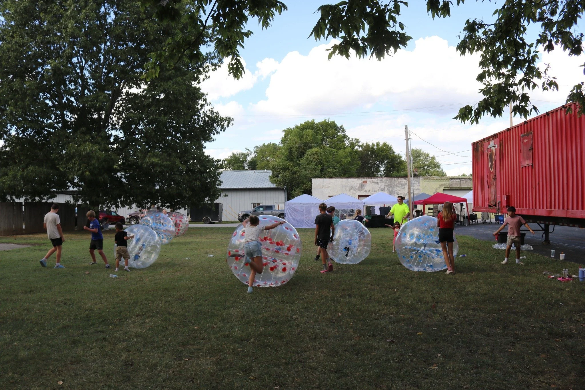 People, including children, playing with large inflatable bubble toys outdoors in a grassy area. There are trees, tents, and a red shipping container in the background.