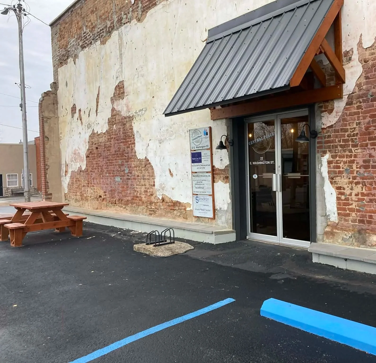 Exterior of a brick building with a weathered wall partially covered in peeling paint, a glass door entrance with a black awning, and a small parking area with blue parking lines and a wooden picnic table.