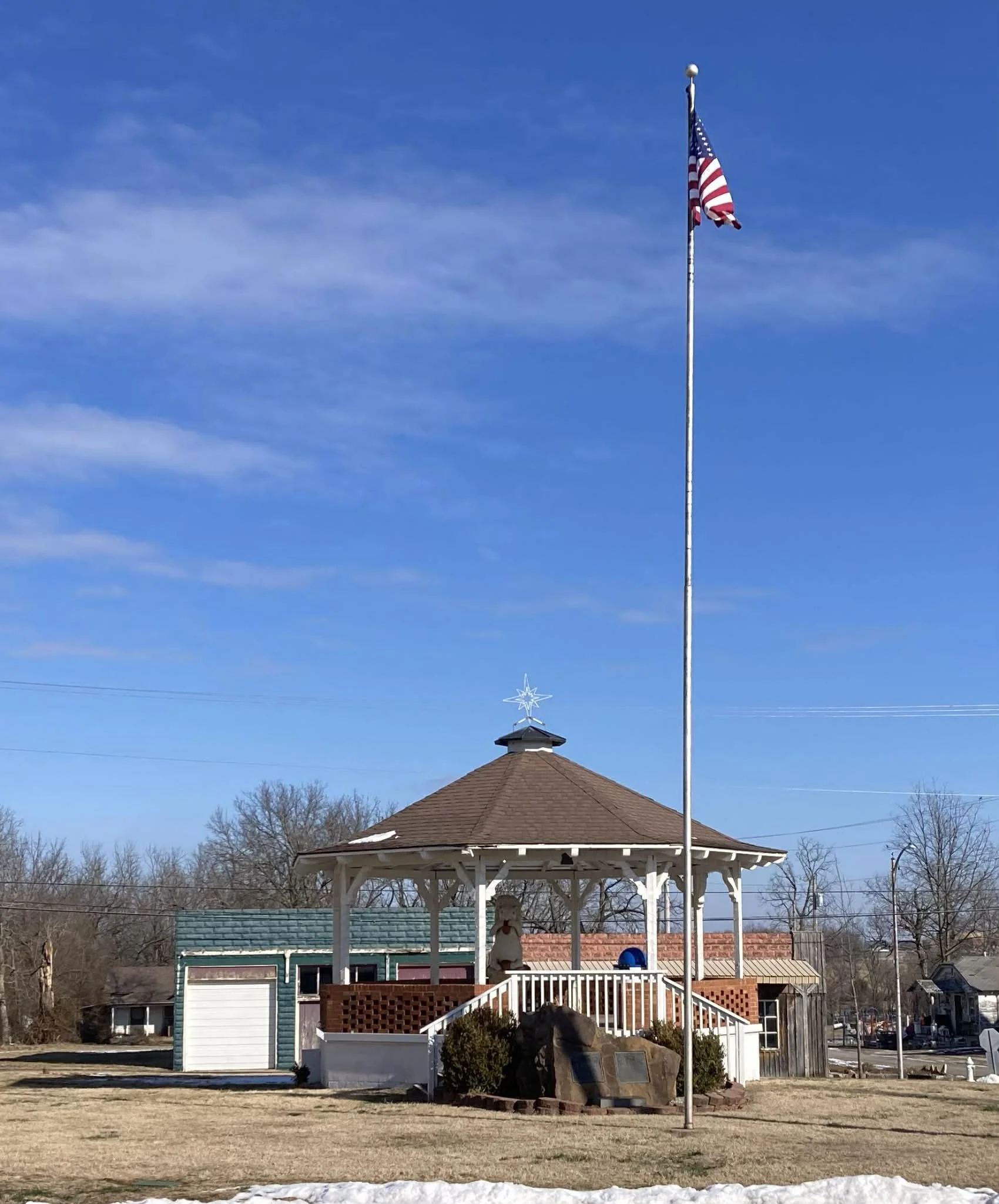 A park with a white gazebo, a tall American flagpole with an American flag, blue sky, leafless trees, and some snow patches on the ground.