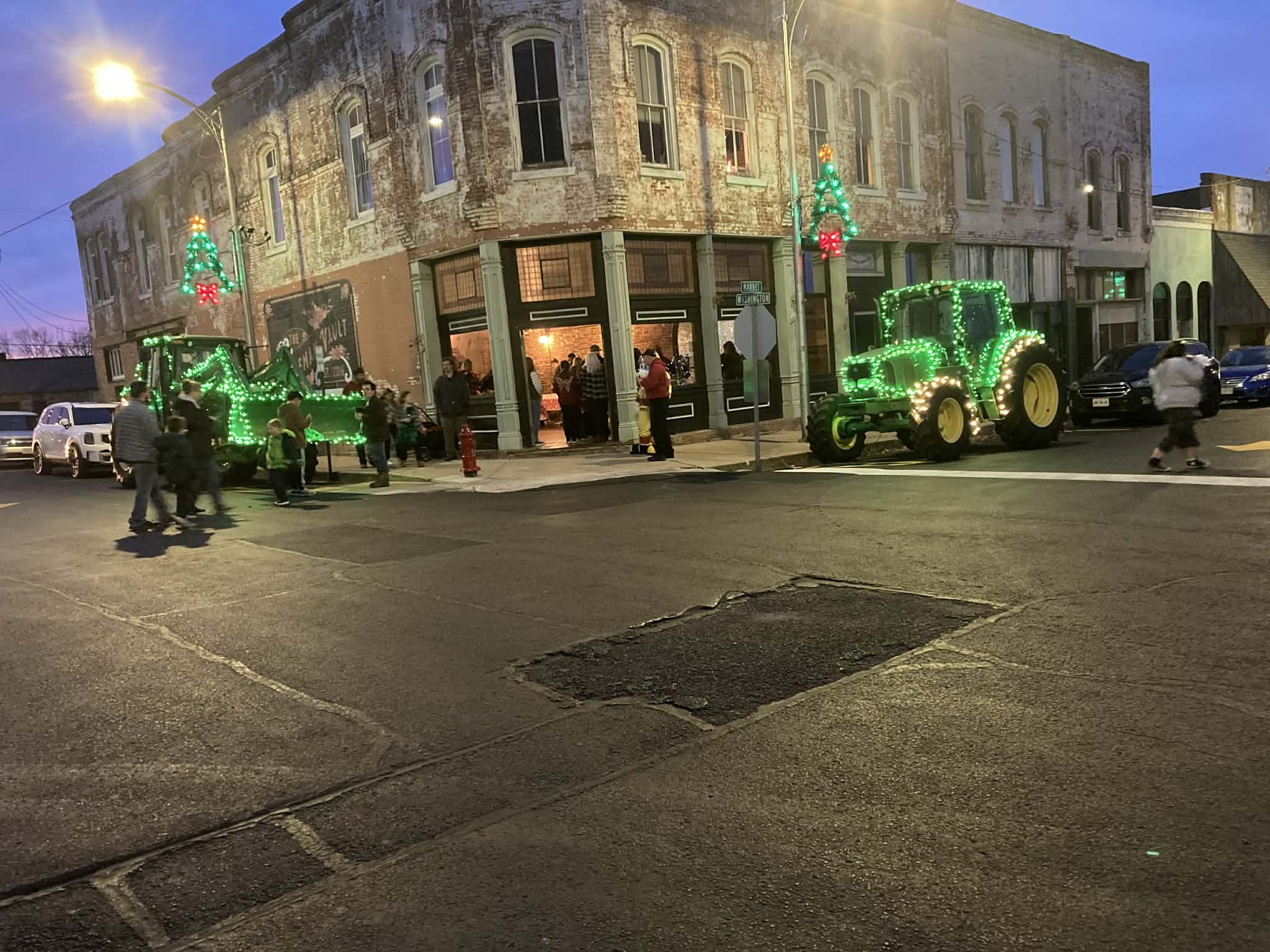 Street scene during evening with decorated green neon Christmas tractor and trailer, and Christmas trees with ornaments, outside a brick building with large windows, some people walking and gathered around, and parked cars.