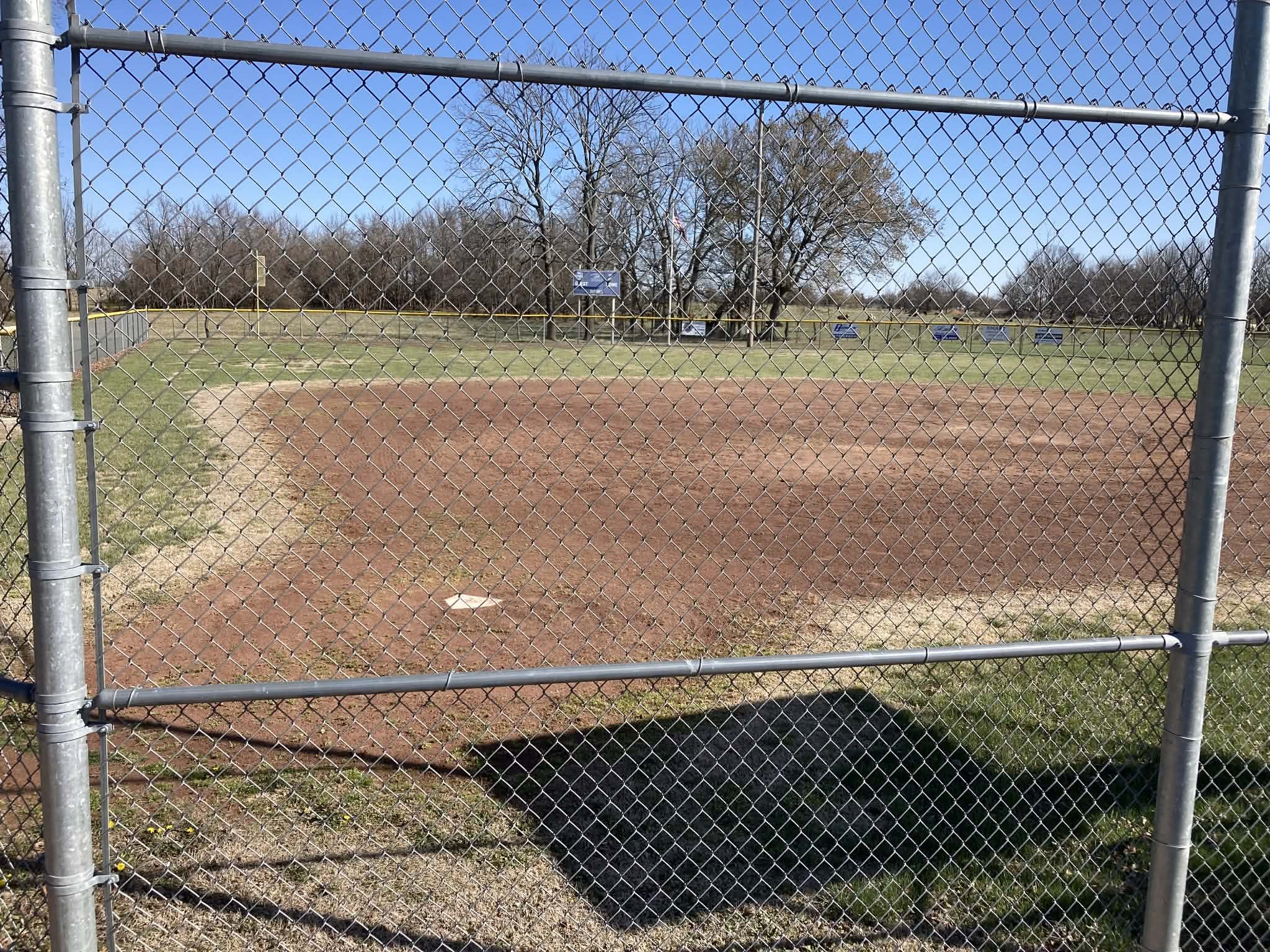 Empty baseball field seen through a chain-link fence, trees in the background, blue sky, grass and dirt infield.