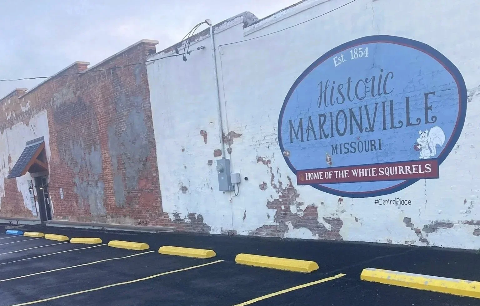 A mural on a brick building wall reads 'Historic Marionville Missouri, Est. 1854, Home of the White Squirrels.' There is a small parking area with yellow painted speed bumps in front of the building.