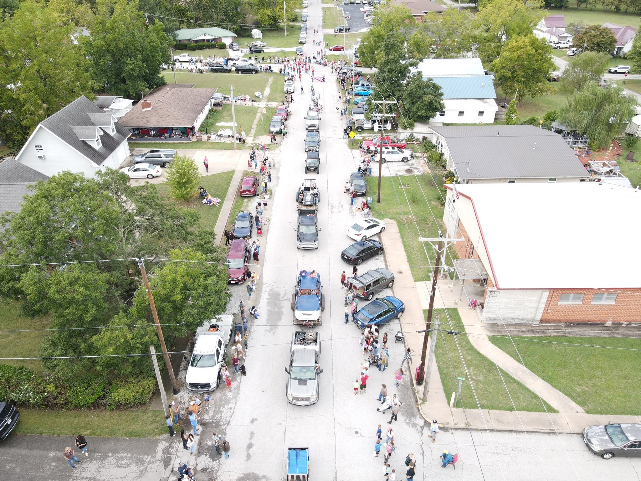 An aerial view of a street during a community event with people walking and shopping along lined-up cars, surrounded by houses and trees.
