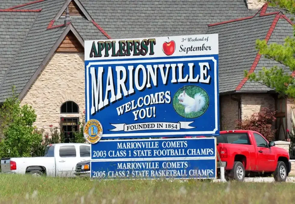 A town sign welcoming visitors to Marionville. The sign features a picture of a white squirrel, mentions an apple festival in September, and highlights local sports achievements from 2003 and 2005.