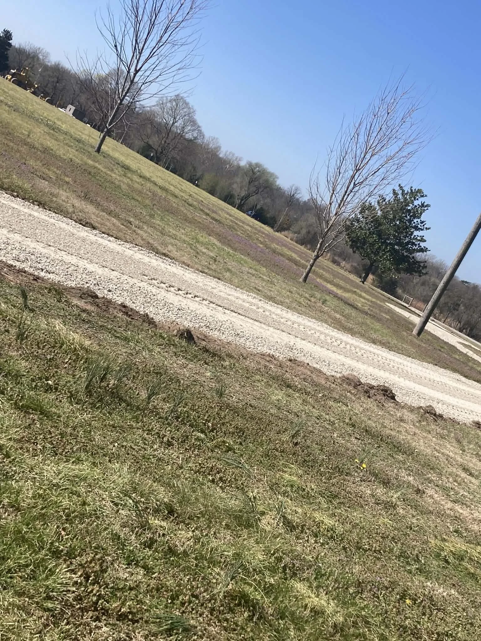 An outdoor park scene with a gravel pathway running through grassy areas, leafless trees, and a clear blue sky.