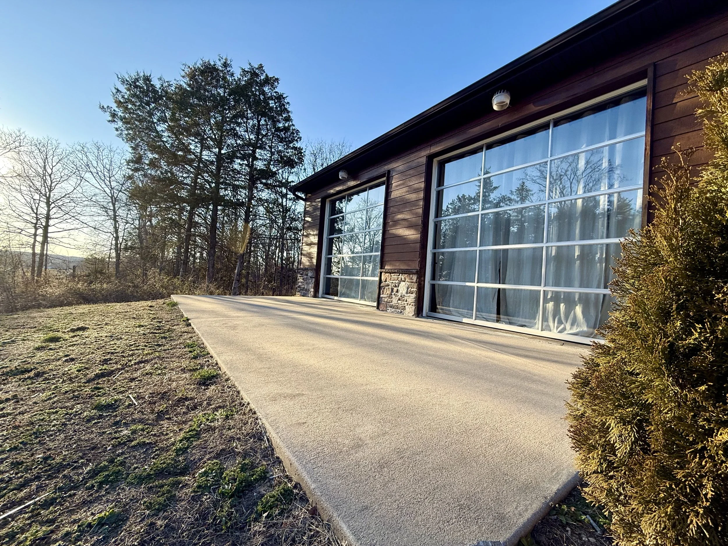 View of a house with large glass windows, a concrete porch, and surrounding trees in the background during daytime.