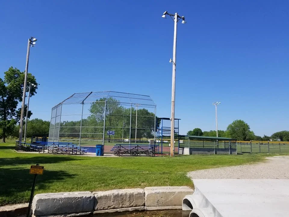 Empty baseball field with chain-link fence, bleachers, and tall light poles under a clear blue sky.