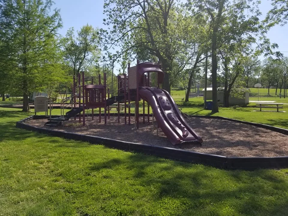 A playground with a purple slide and climbing structure in a park with green grass and trees.