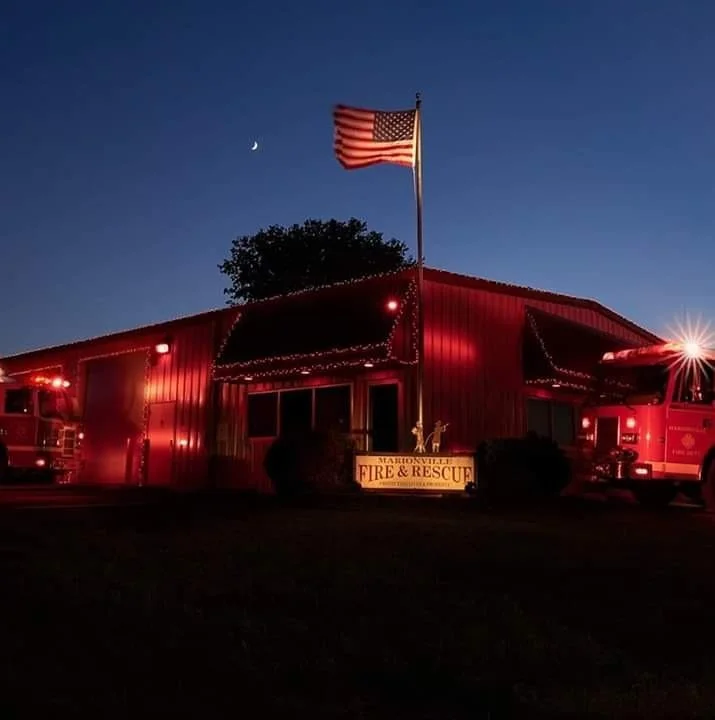 Nighttime image of a fire station with red lights, an American flag on a flagpole, and a sign that reads 'Marionville Fire & Rescue'.