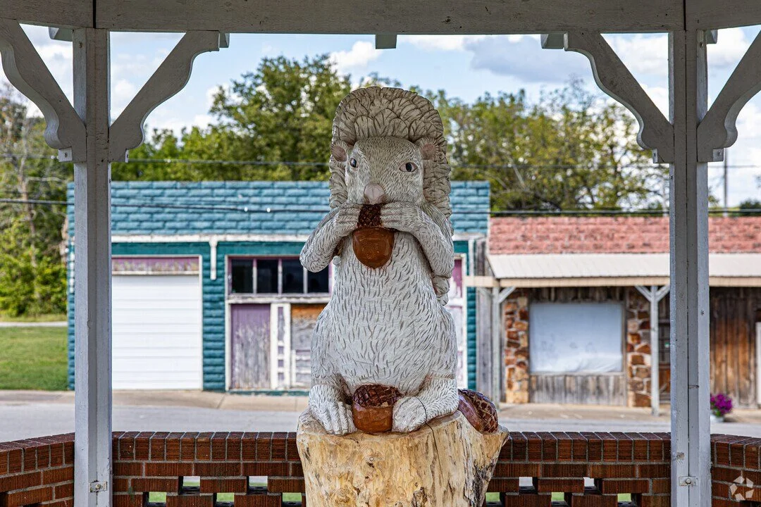 A statue of a squirrel holding an acorn, sitting on a tree stump in an outdoor setting with buildings and trees in the background.