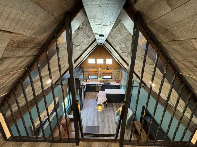 View from a loft looking down into a modern, rustic-style living room and kitchen area with wooden walls, high ceiling, and small windows.