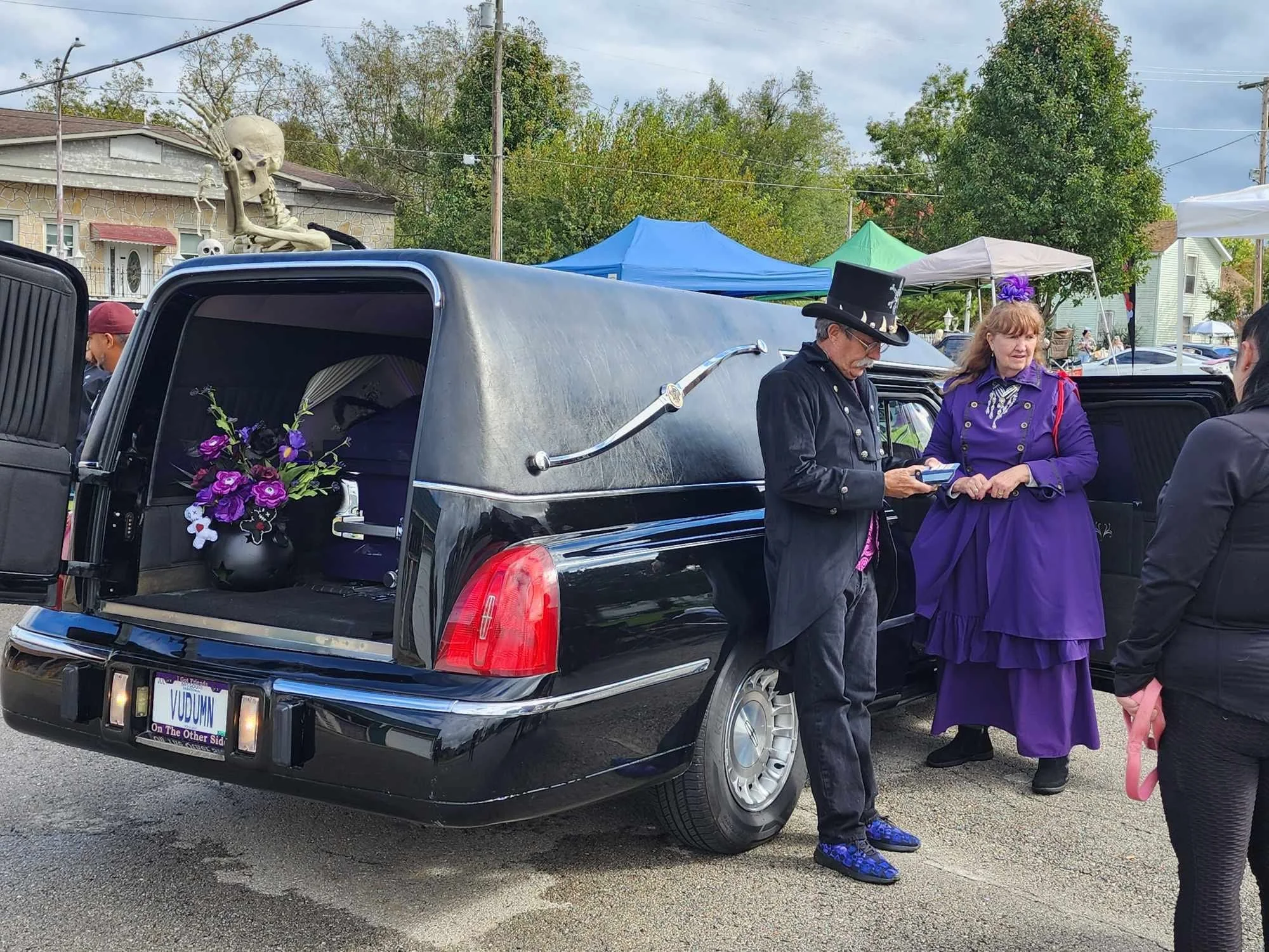 A black hearse with a purple floral arrangement in the back, parked at an outdoor event with people dressed in gothic and steampunk attire. A skeleton decoration is on top of the hearse's back end.