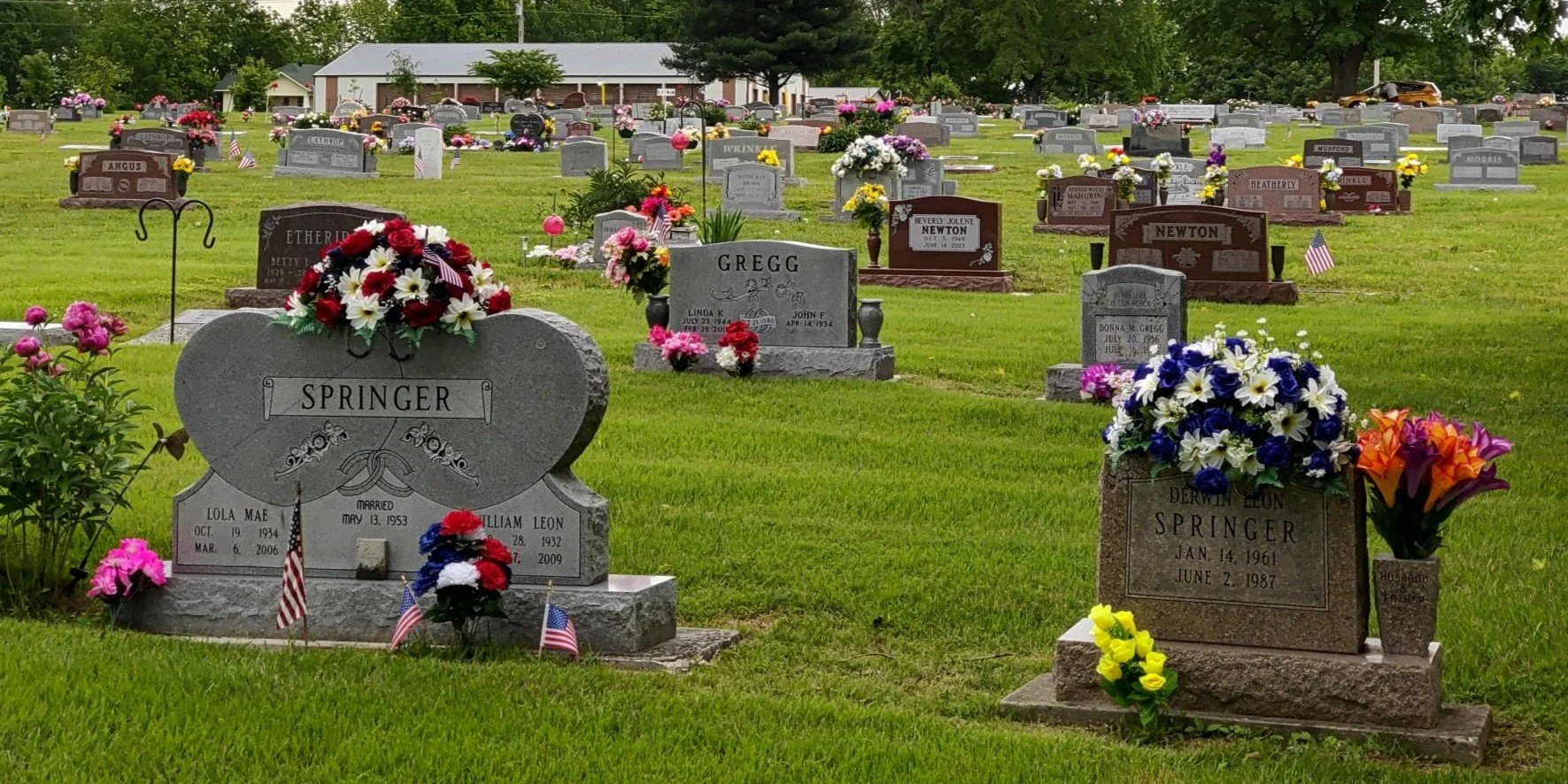 A cemetery with rows of gravestones decorated with colorful flowers and small American flags, and a farmhouse or barn in the background.