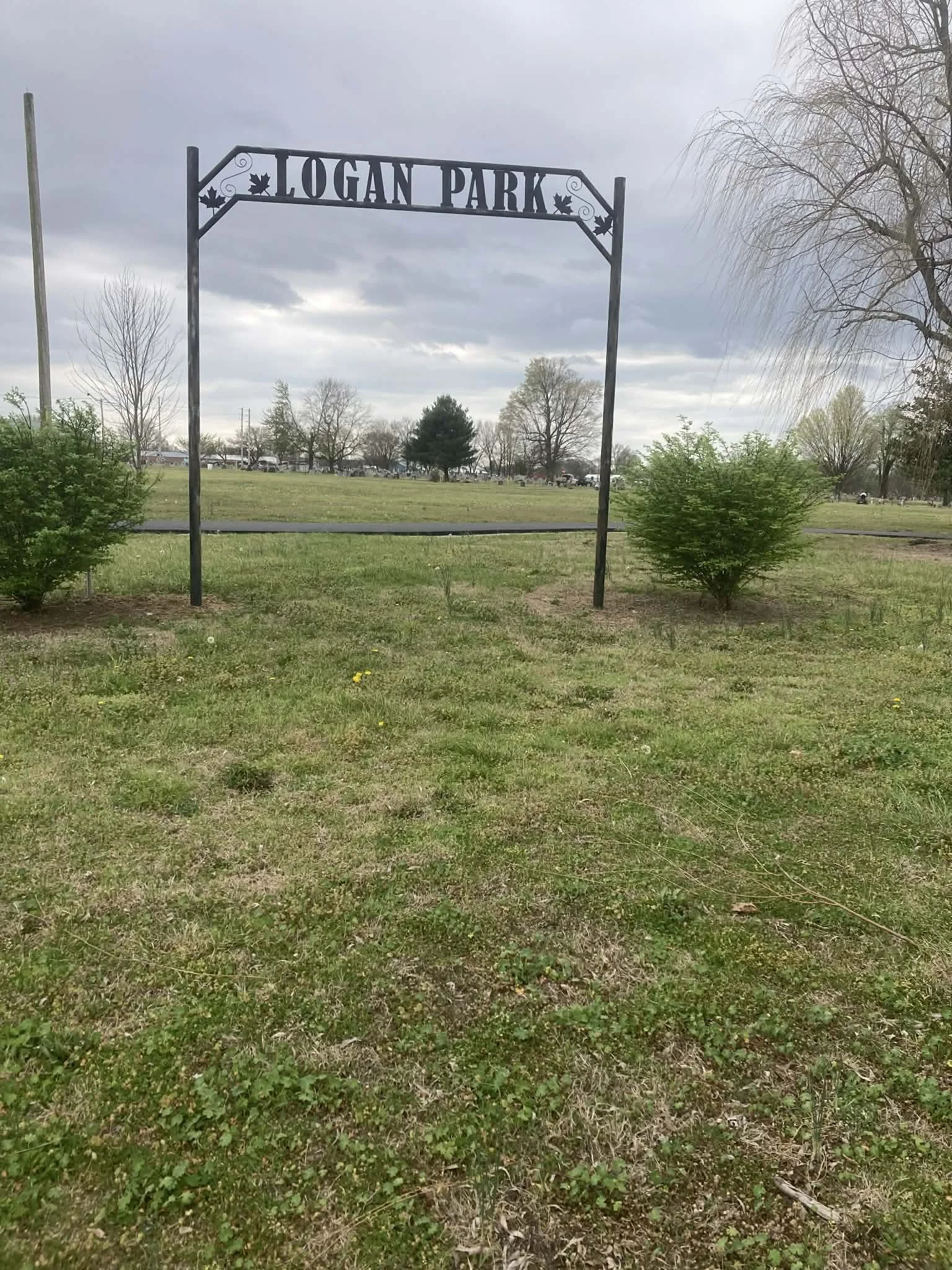Entrance sign for Logan Park with grass, trees, and cloudy sky in the background.