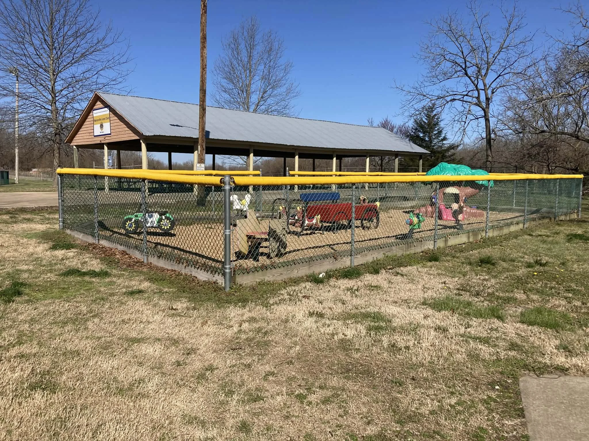 A fenced children's playground with various playground equipment and a covered shelter, located in a park with trees and a clear blue sky. Marionville, MO city park.