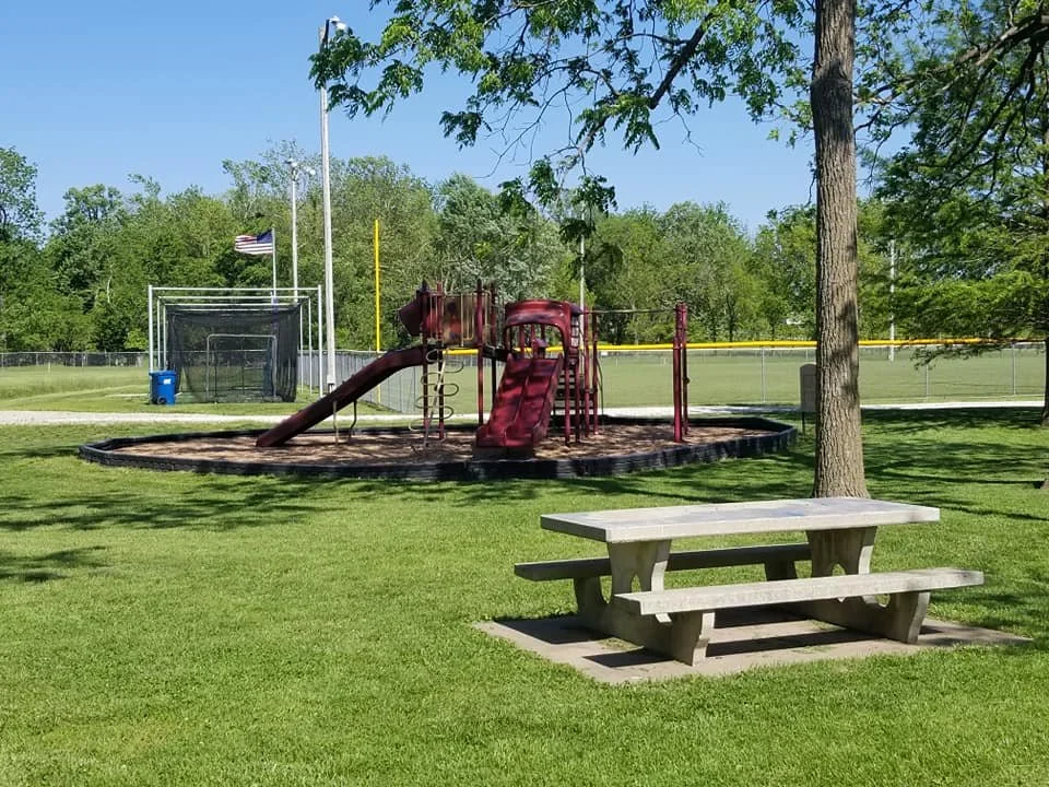 A playground with a red slide and climbing structure surrounded by a sandpit, a picnic table, trees, a football field, and an American flag in the background on a sunny day.