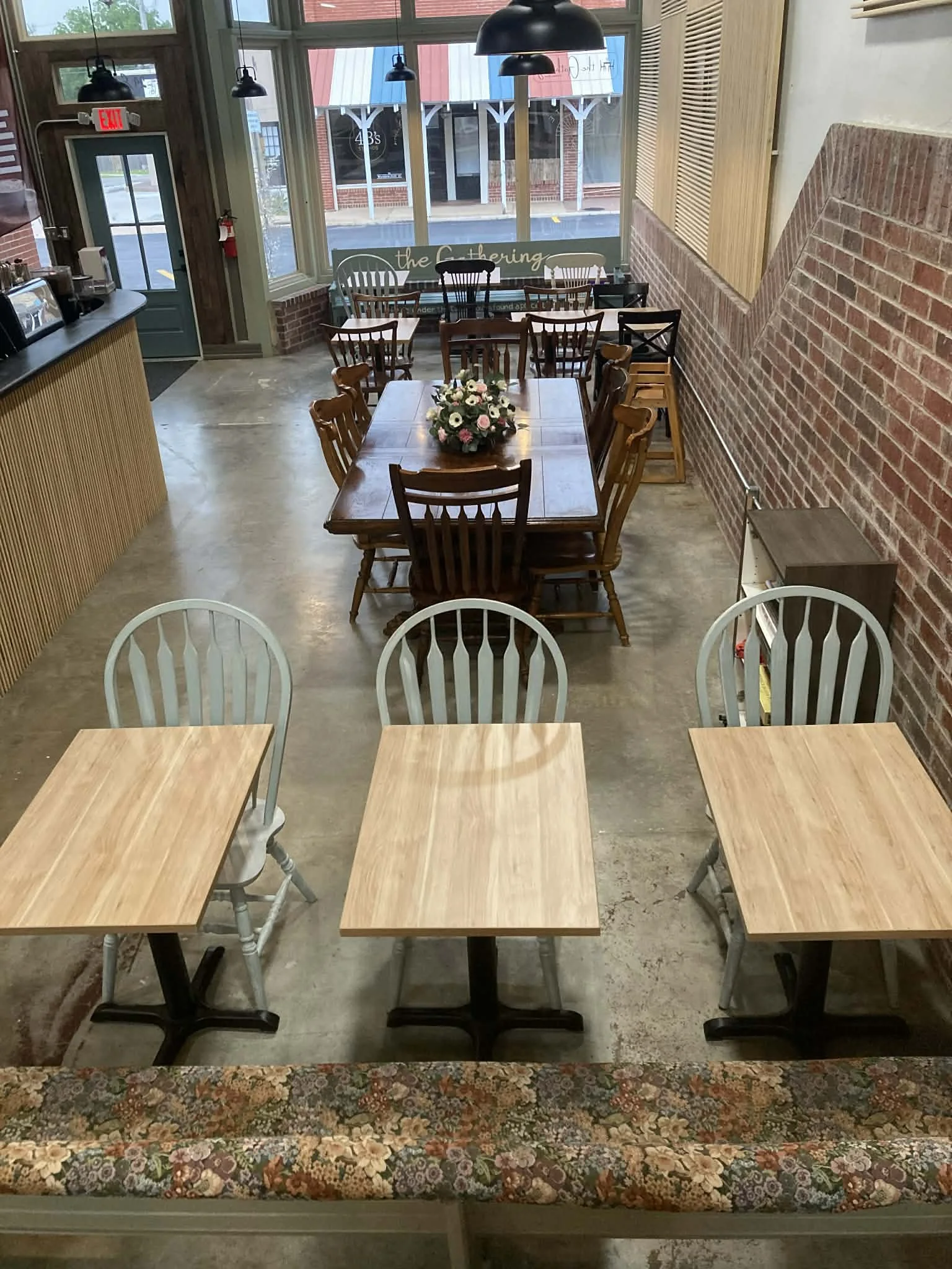 Interior view of a cozy coffee shop with wooden tables and chairs, floral-patterned cushioned seating, brick walls, large windows, and pendant lighting.