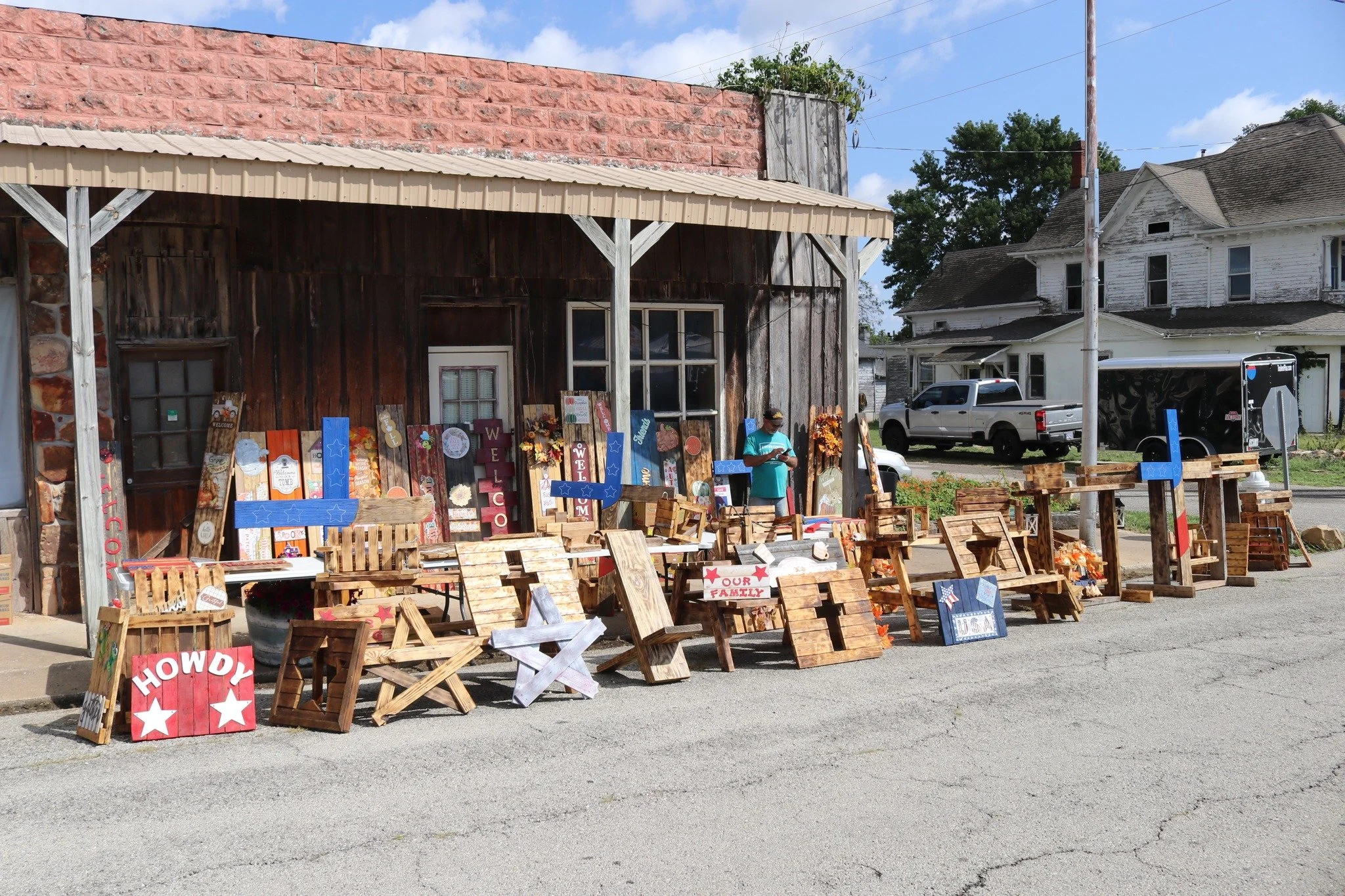 Outdoor display of wooden signs and crafts on a sidewalk outside a rustic building with a wood and stone facade, in a small town setting with houses and vehicles in the background.