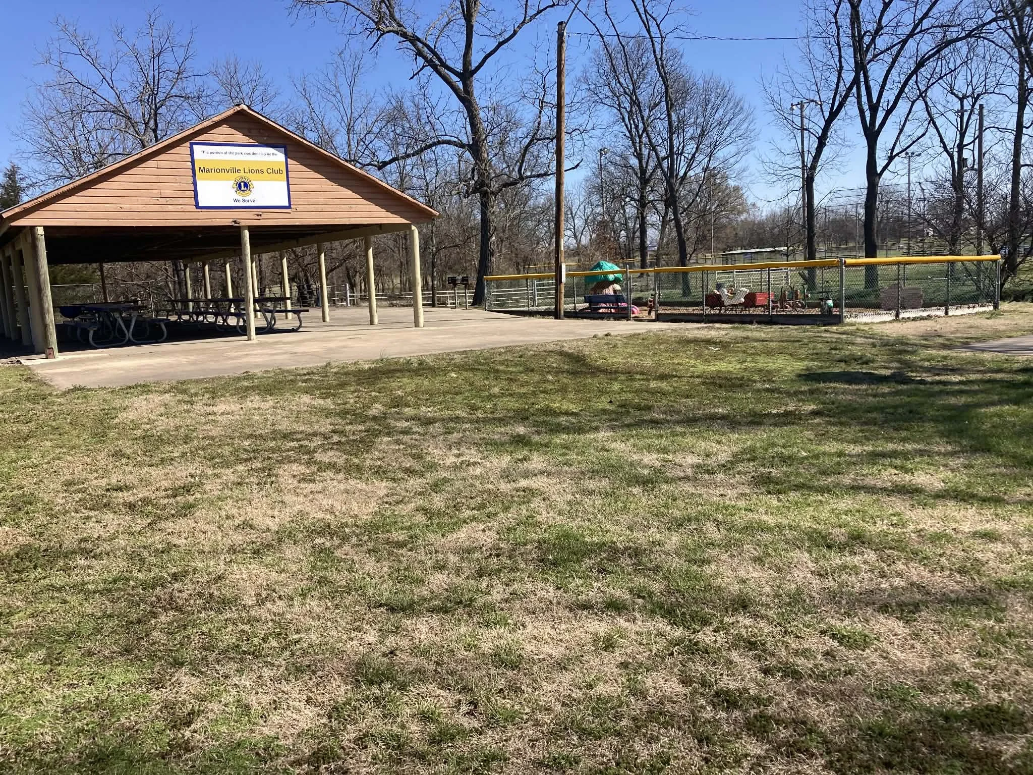 Park with picnic shelter, fenced play area, and leafless trees under a clear blue sky. Marionville, MO city park.