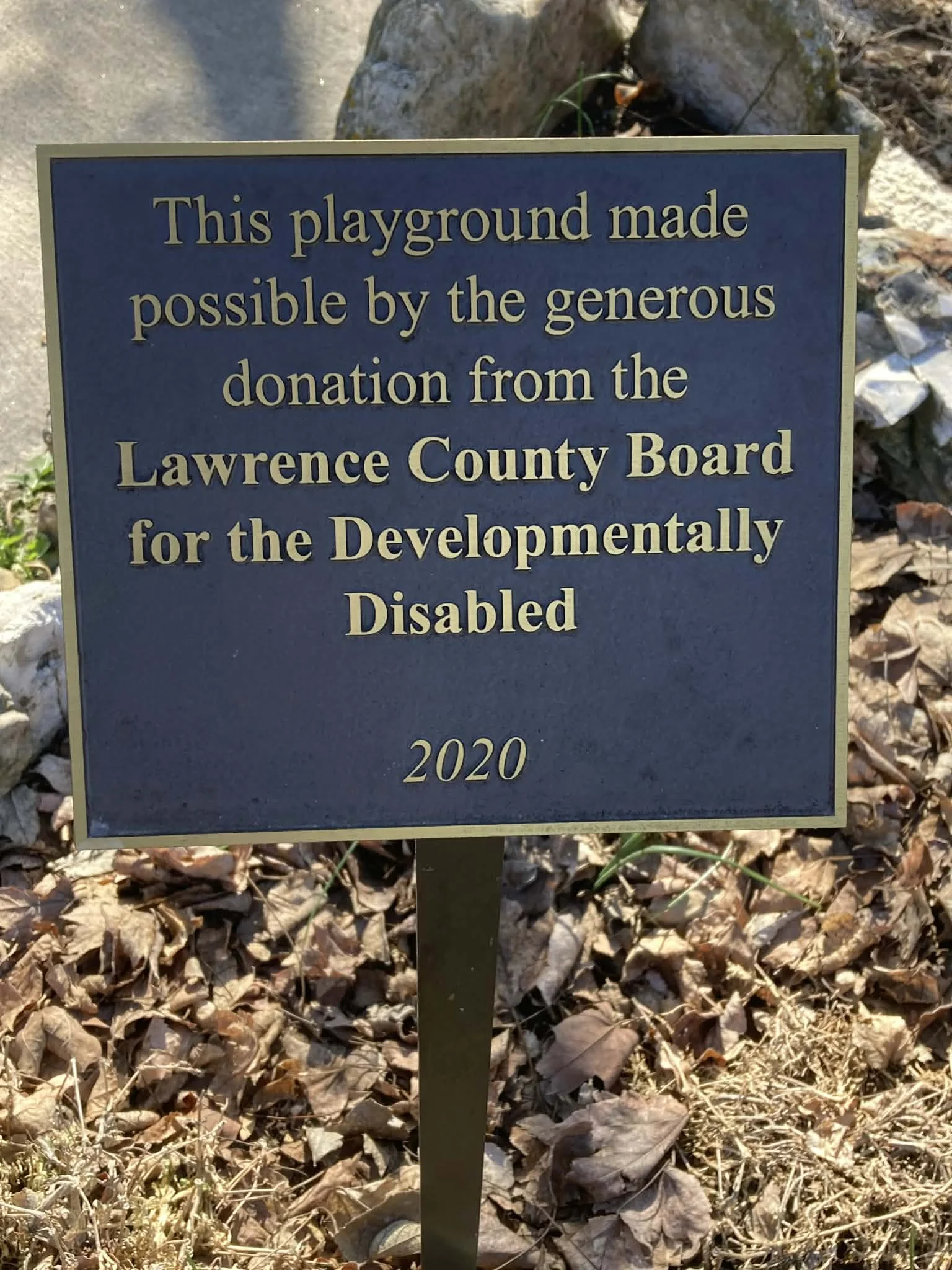A sign indicating the playground was funded by a donation from the Lawrence County Board for the Developmentally Disabled, dated 2020, with fallen leaves and rocks visible around it. Marionville, MO City Park