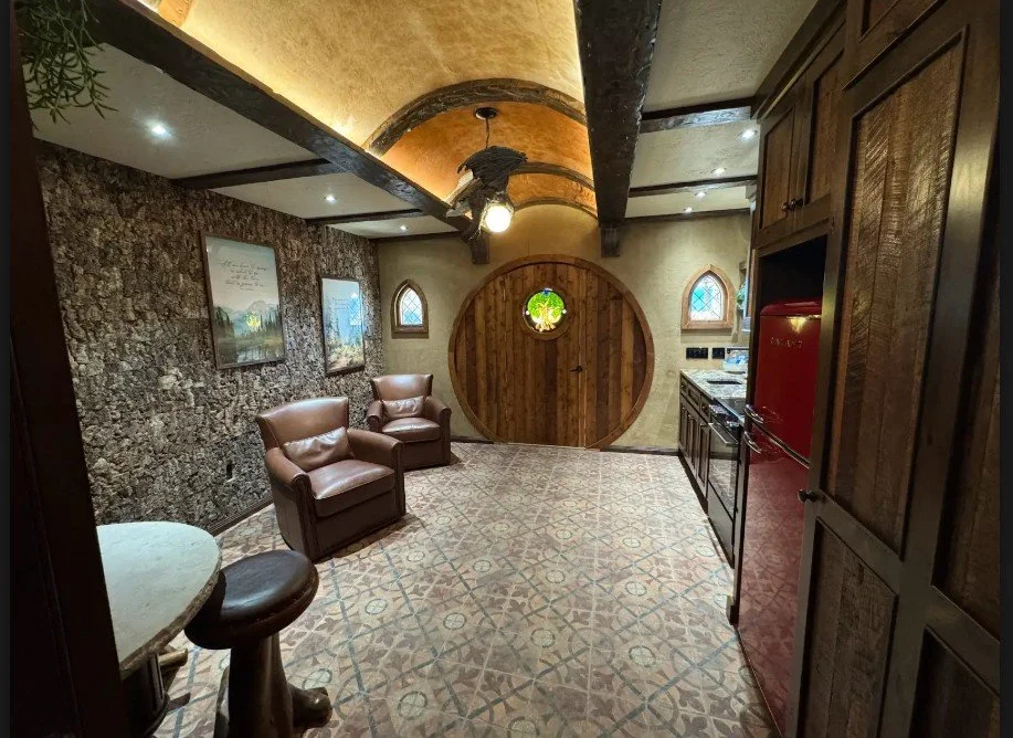 Cozy kitchen with stone and wood decor, round wooden door, two small arched windows, and a seating area with two leather chairs.
