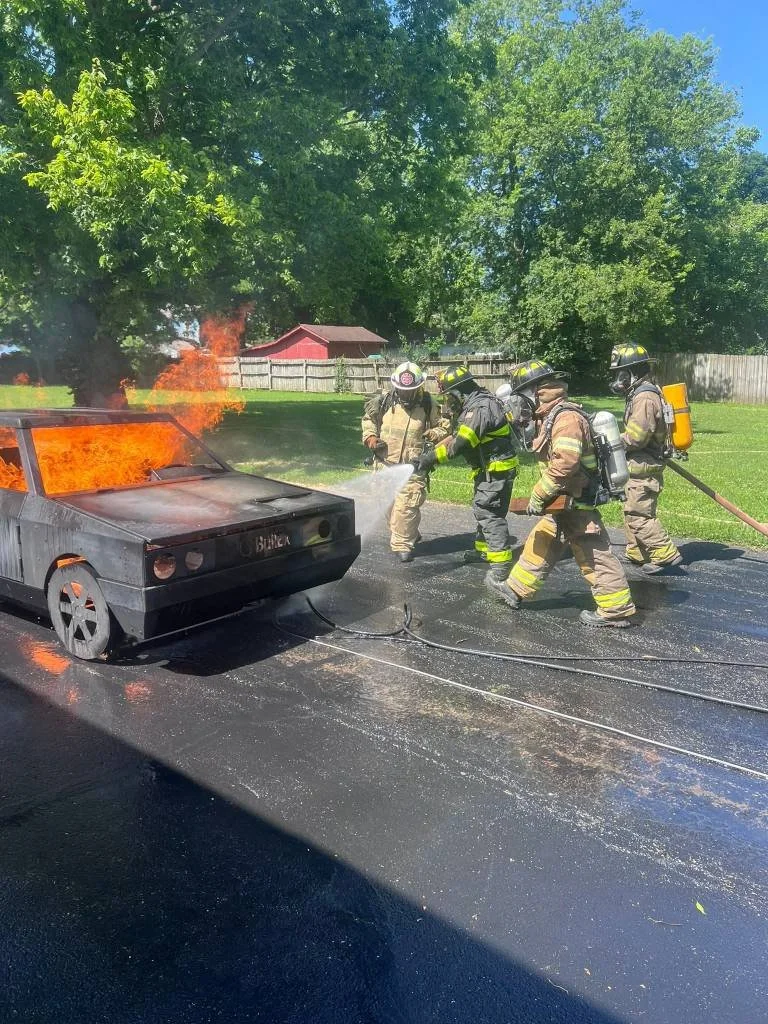 Firefighters extinguishing a car fire on a street, with flames visible on the car's hood and front area, while standing on the blackened asphalt in a residential neighborhood with green trees and a wooden fence.