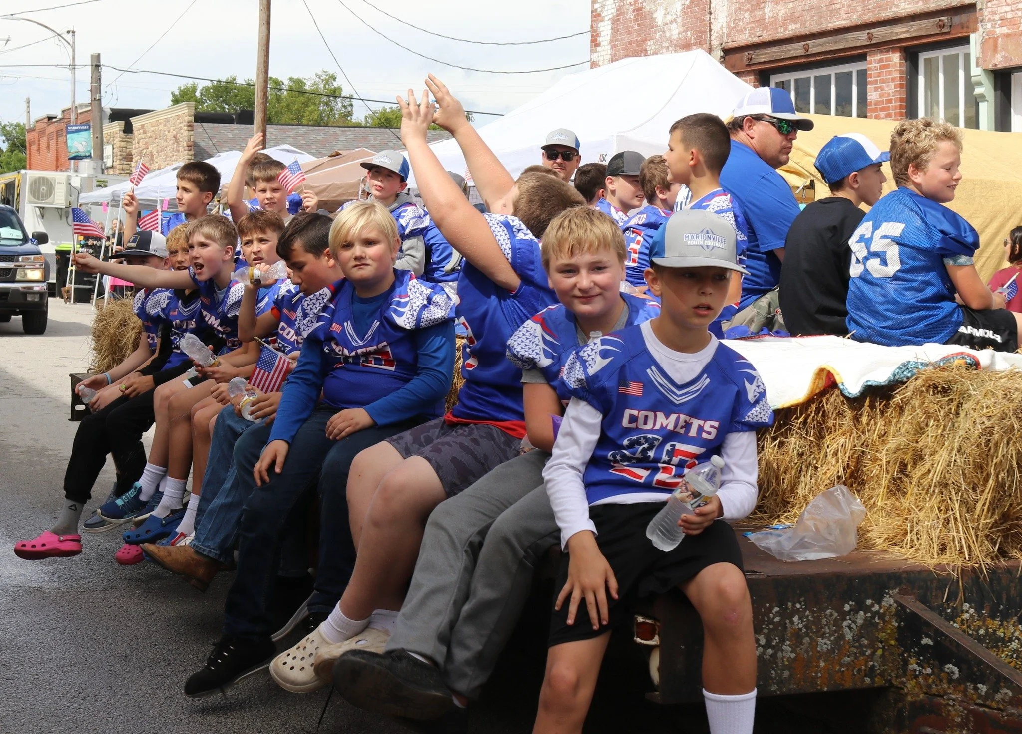 Children wearing blue sports jerseys sitting on a float decorated with hay bales during a parade, someholding small American flags.