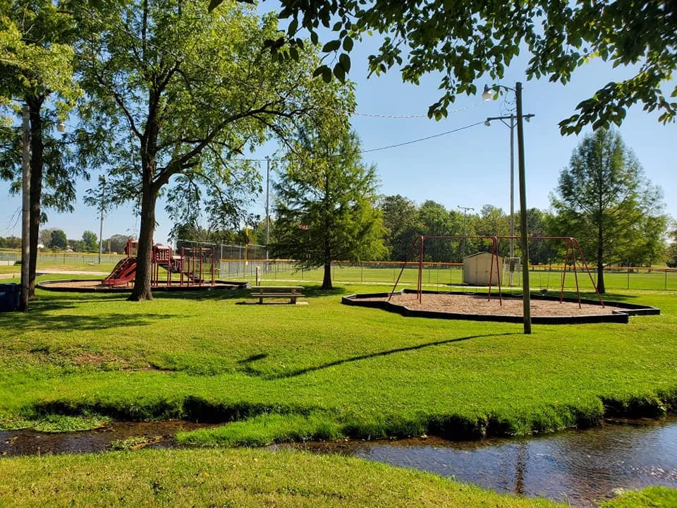 A park with a playground, swings, and a slide, surrounded by trees and grass, with a small stream in the foreground and a blue sky.