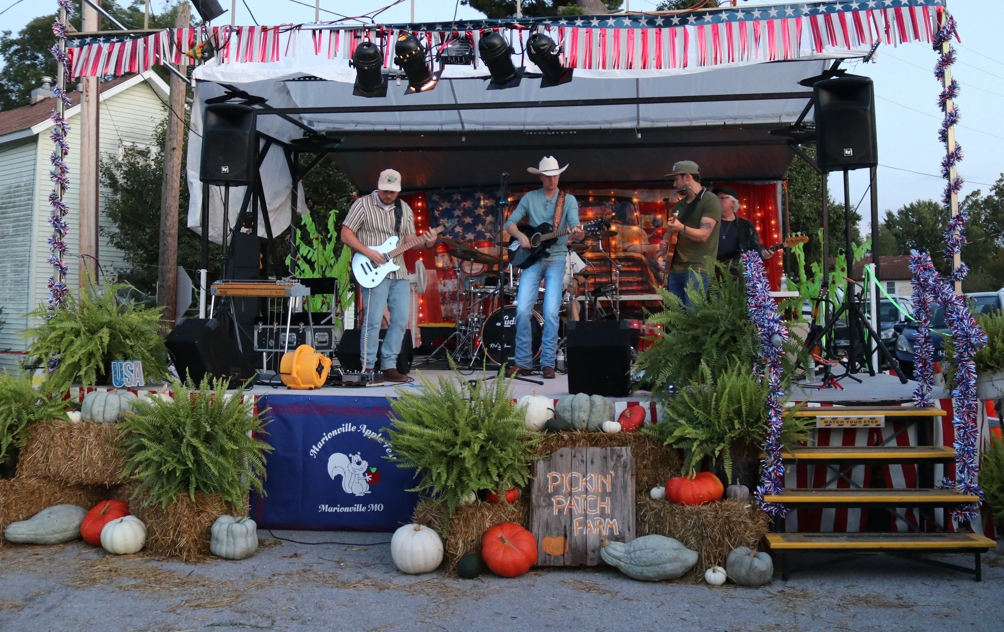 A live band performing on a decorated outdoor stage at a fall festival, with pumpkins and fall decor in front, under an American-themed banner.