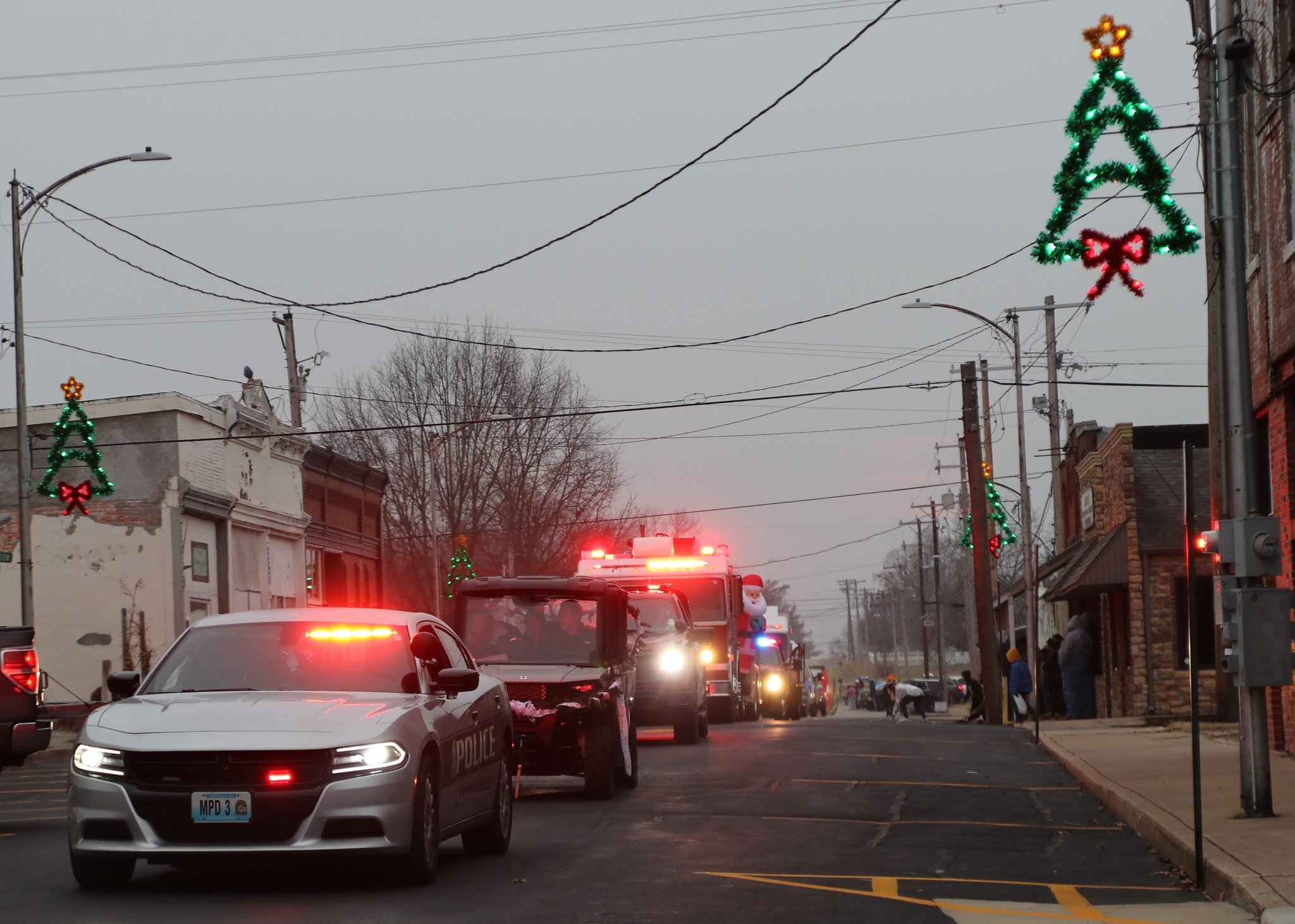 A street scene with police and emergency vehicles, decorated with Christmas lights and ornaments, and people on the sidewalk.