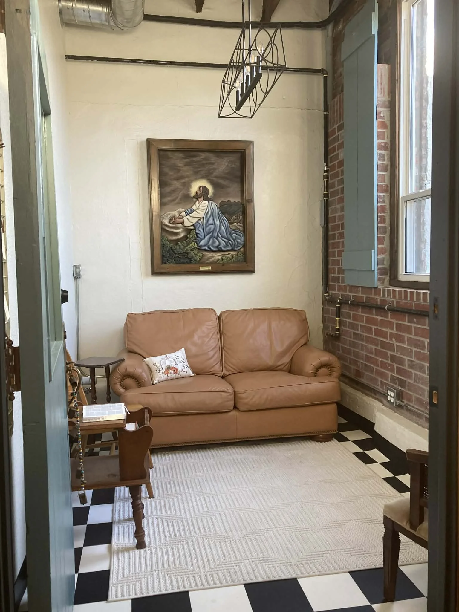 A cozy sitting area with a tan leather sofa, a small embroidered pillow, a wooden chair, a white rug, a window with green shutters, a framed religious painting, exposed brick wall, and a black-and-white checkered floor.