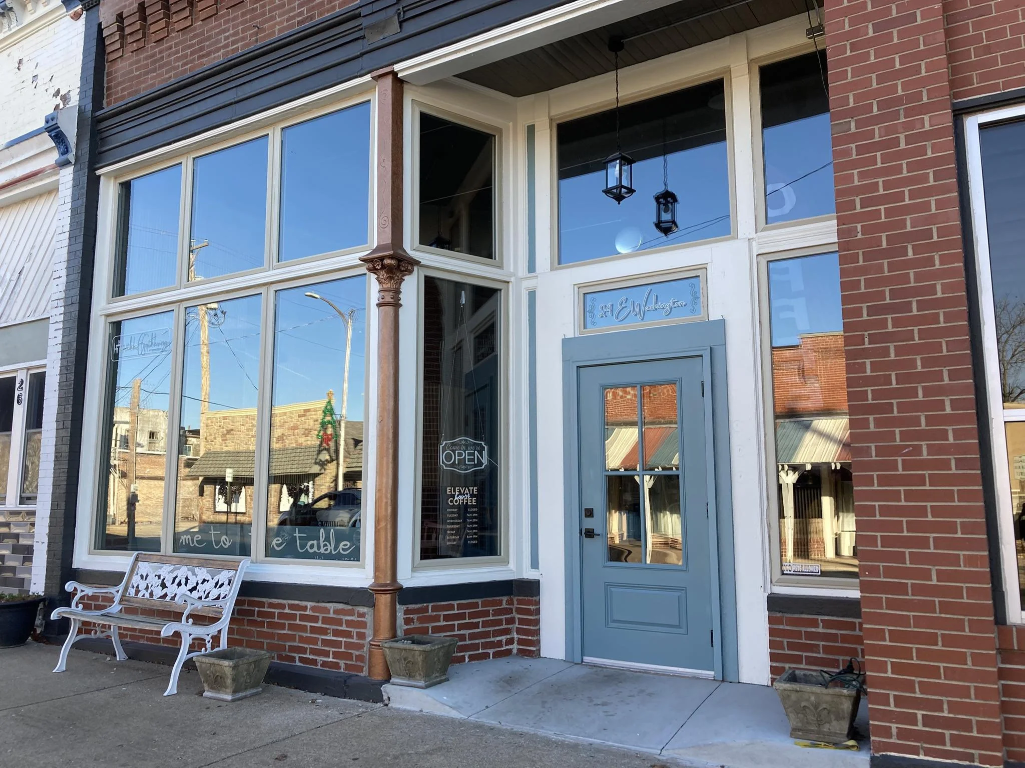 Exterior of a coffee shop with a blue door, large window panes, and a white decorative bench outside. There are two empty planters on the sidewalk, and the shop has a sign indicating it is open.