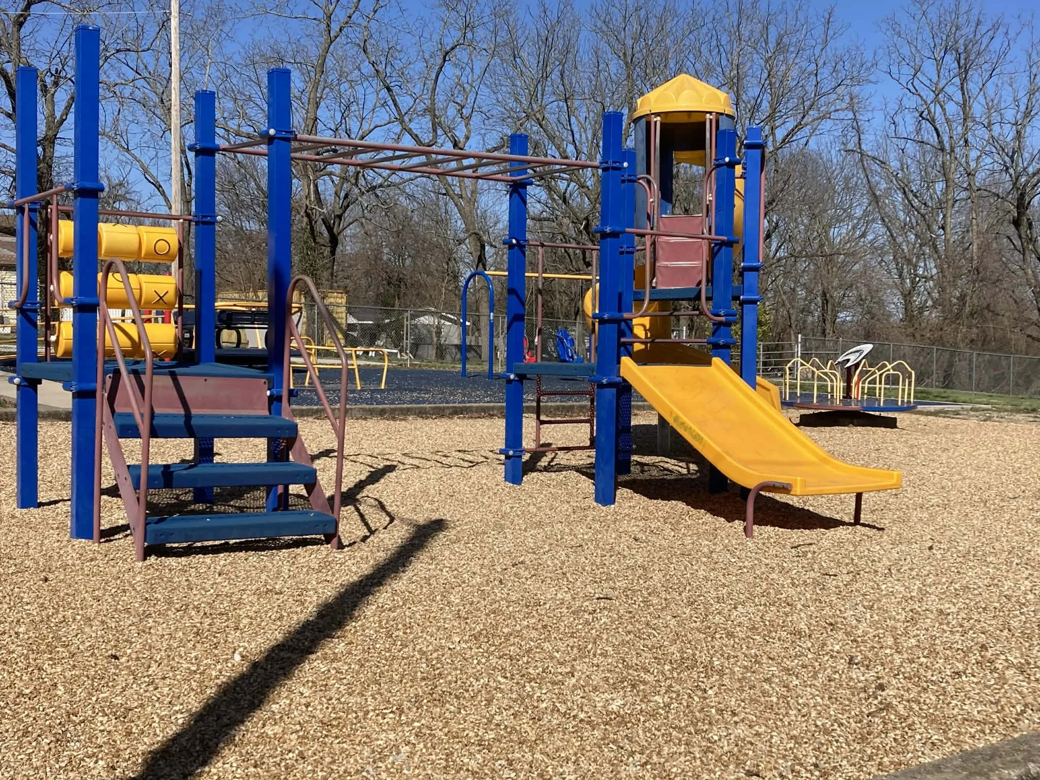 Colorful playground equipment, including a yellow slide, blue and brown structures, and climbing features, in a park with bare trees and a chain-link fence in the background. Marionville, MO City Park
